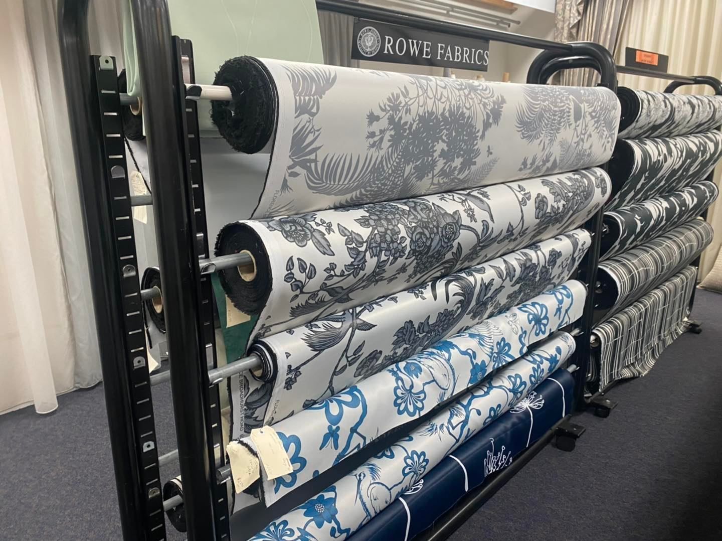 A Metal Display Rack Holds Multiple Rolls Of Patterned Fabric In Shades Of White, Gray, And Blue — Instyle Curtains & Blinds in Bungalow, QLD