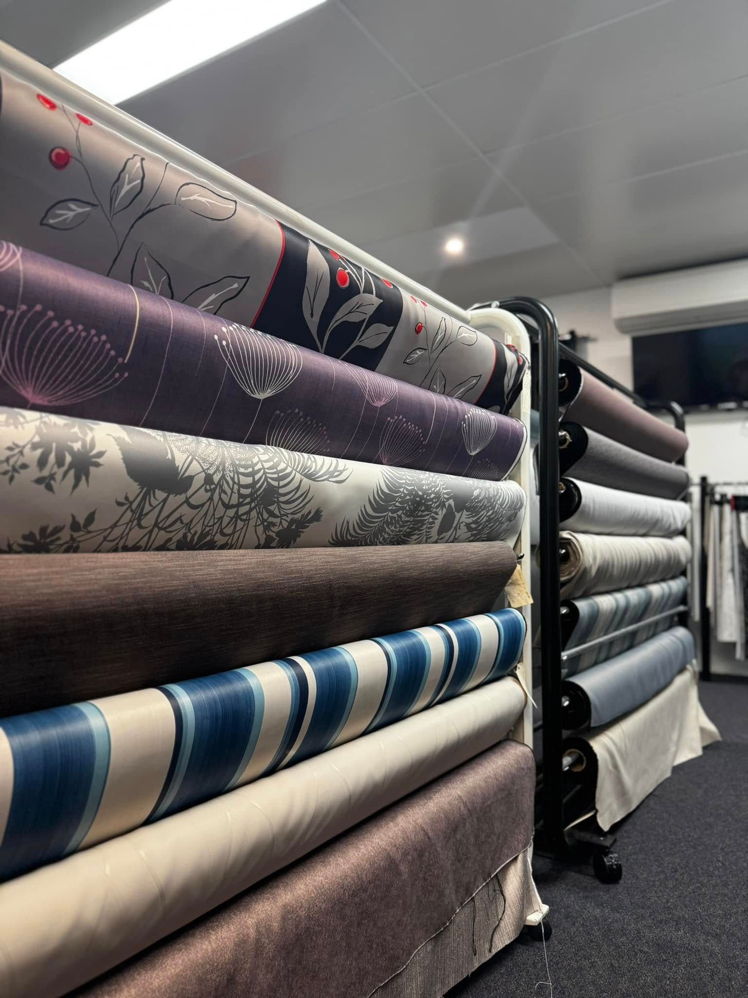 A Fabric Shop Rack With Multiple Rolls Of Patterned And Textured Textiles In Shades Of Blue, Grey, And Brown — Instyle Curtains & Blinds in Bungalow, QLD