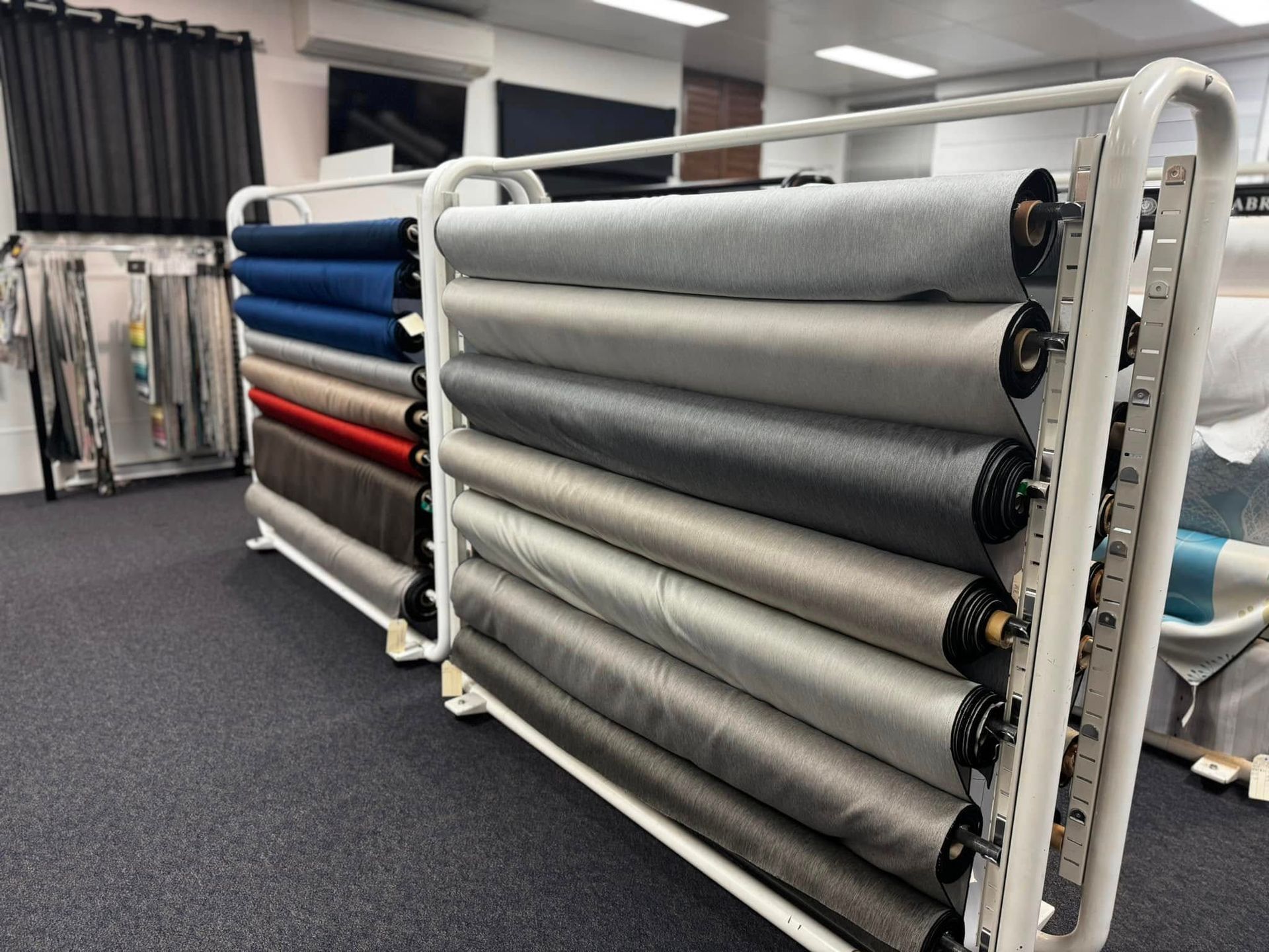 Two Tiered Metal Racks Holding Rolls Of Fabric In Various Shades Of Grey, Blue, And Red Inside A Retail Store — Instyle Curtains & Blinds in Bungalow, QLD