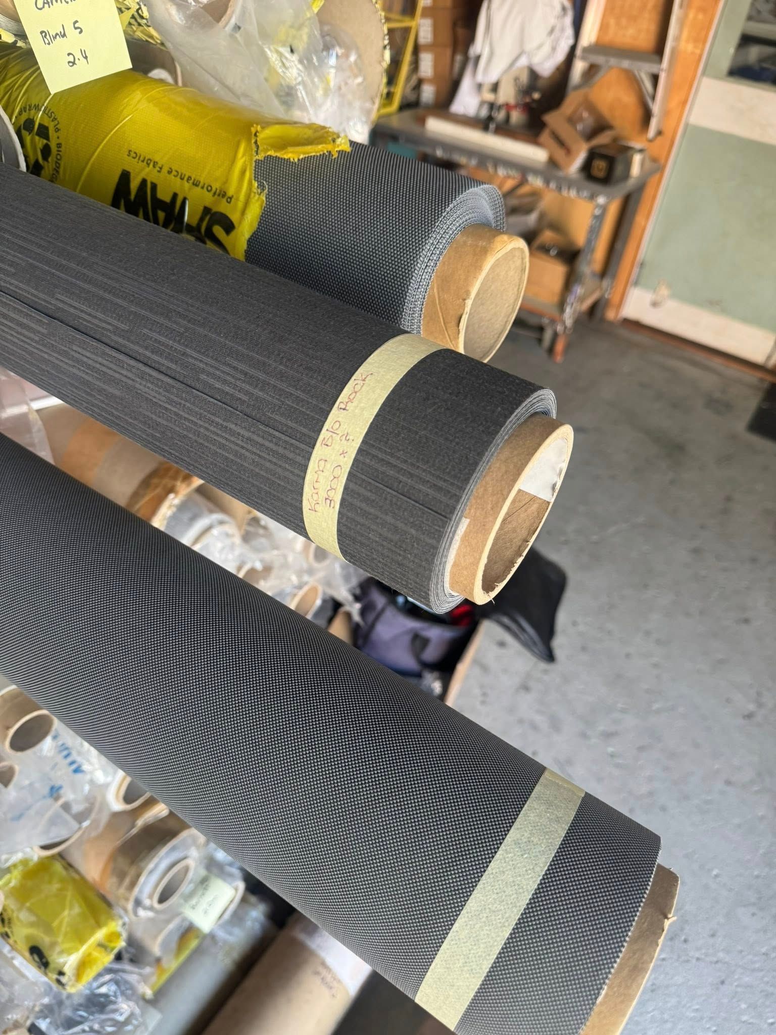 Rolls Of Dark, Textured Screen Or Fabric Material Are Stacked On Shelving In A Storage Area — Instyle Curtains & Blinds in Bungalow, QLD