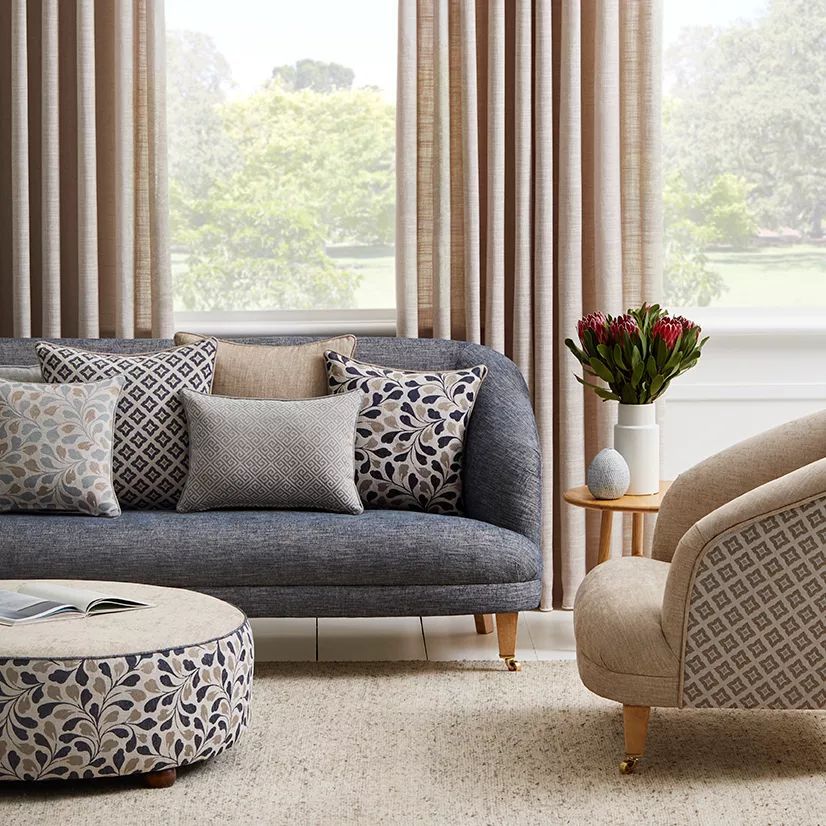 A grey sofa with patterned and solid cushions, a round patterned ottoman, and a beige armchair in a sunlit living room.