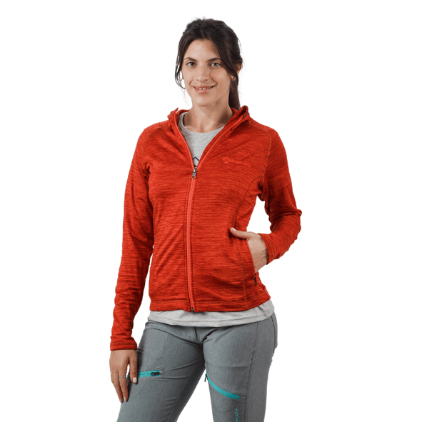 Mujer con chaqueta roja con capucha y pantalones grises, de pie con la mano en el bolsillo, sonriendo.