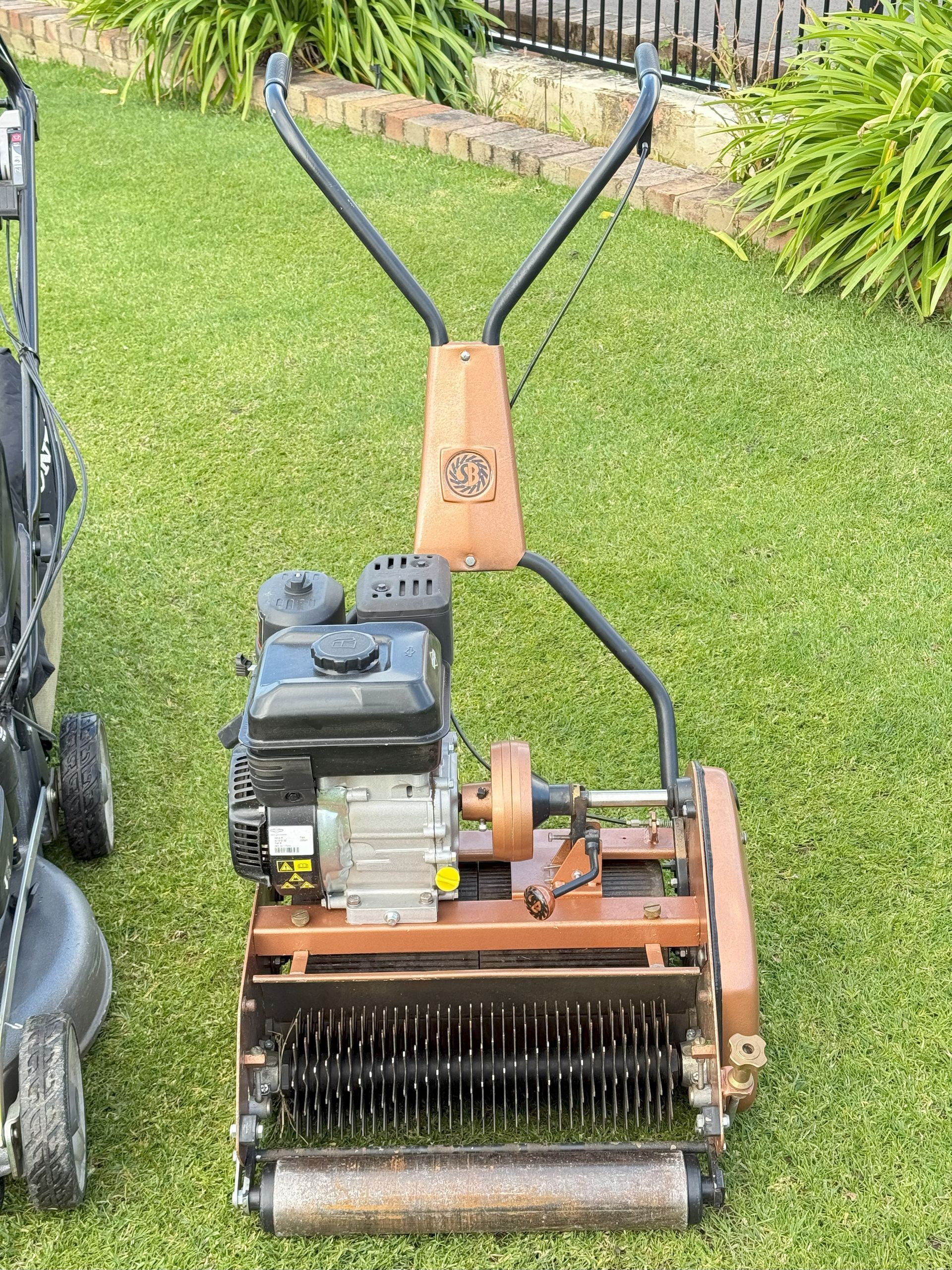 A walk-behind, reel-type lawn mower on a green lawn. It has a brown frame and handles, with a black engine and cutting reel.