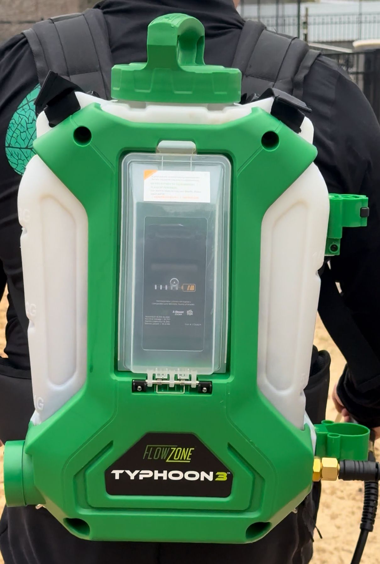 Back view of a person wearing a green and white Typhoon 3 backpack sprayer. The sprayer has a battery compartment and a carrying handle.
