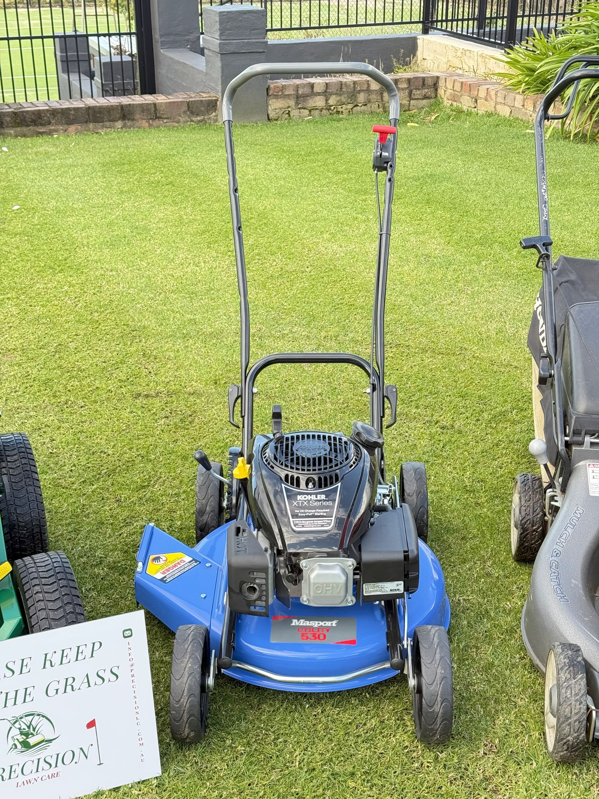 Blue lawnmower on grass, standing between two other mowers. Sunlight illuminates the scene outdoors.