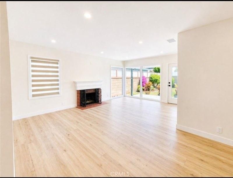 Bright empty living room with light wood floors, white walls, fireplace, and sliding glass doors to a patio.