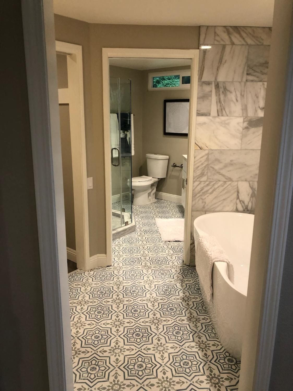 Narrow bathroom hallway with patterned tile floor, marble walls, bathtub, toilet, and glass shower door.