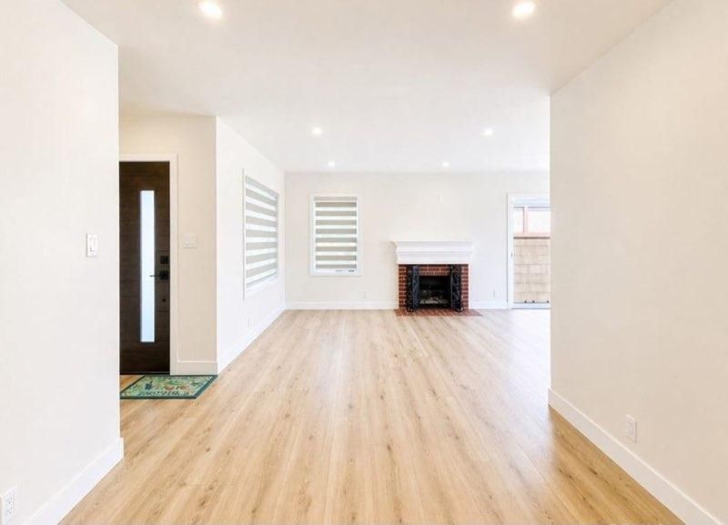 Bright empty living room with light wood floors, white walls, and a fireplace at the far end.