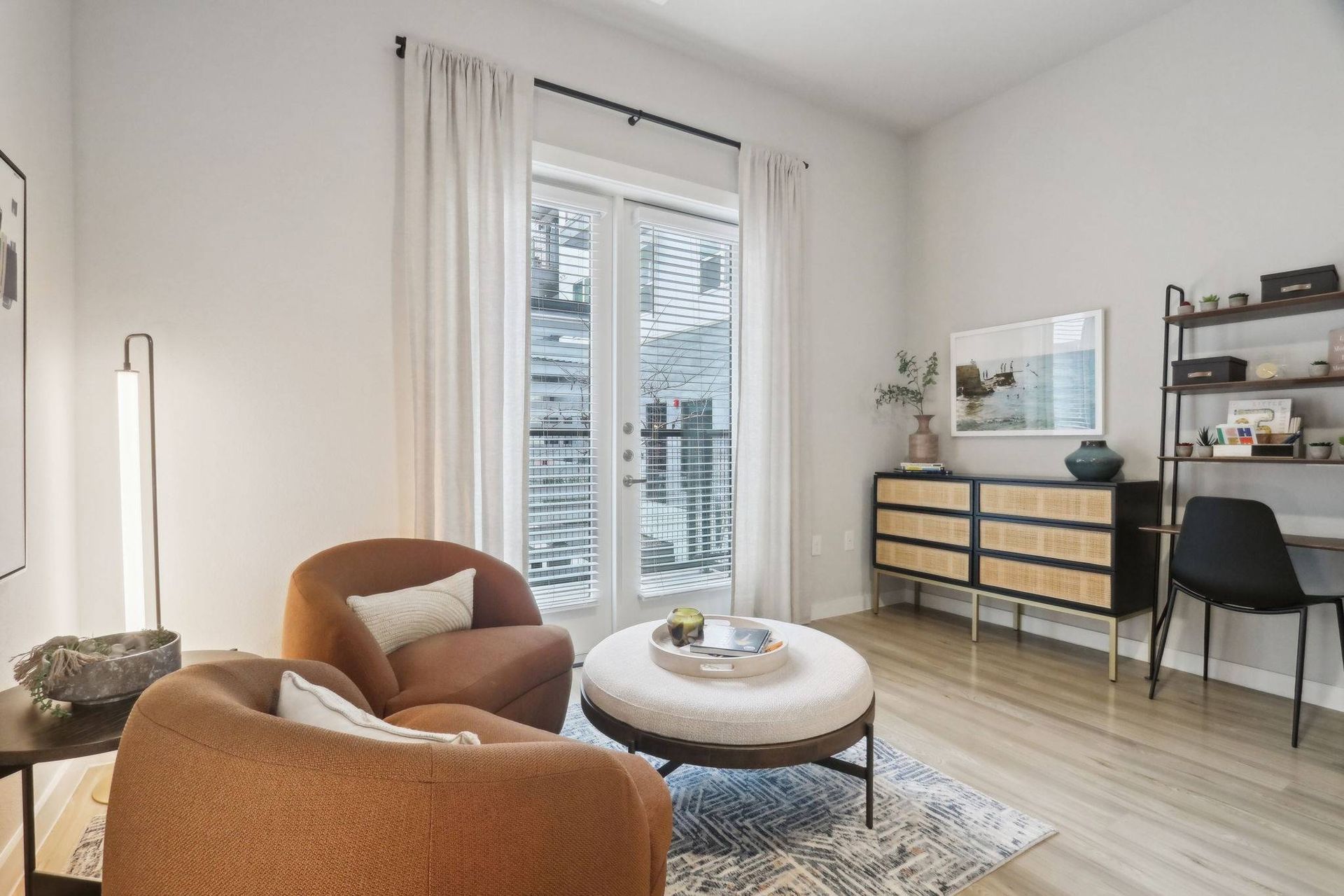 Living room in an apartment with orange chairs, a round ottoman, and a desk against a wall.
