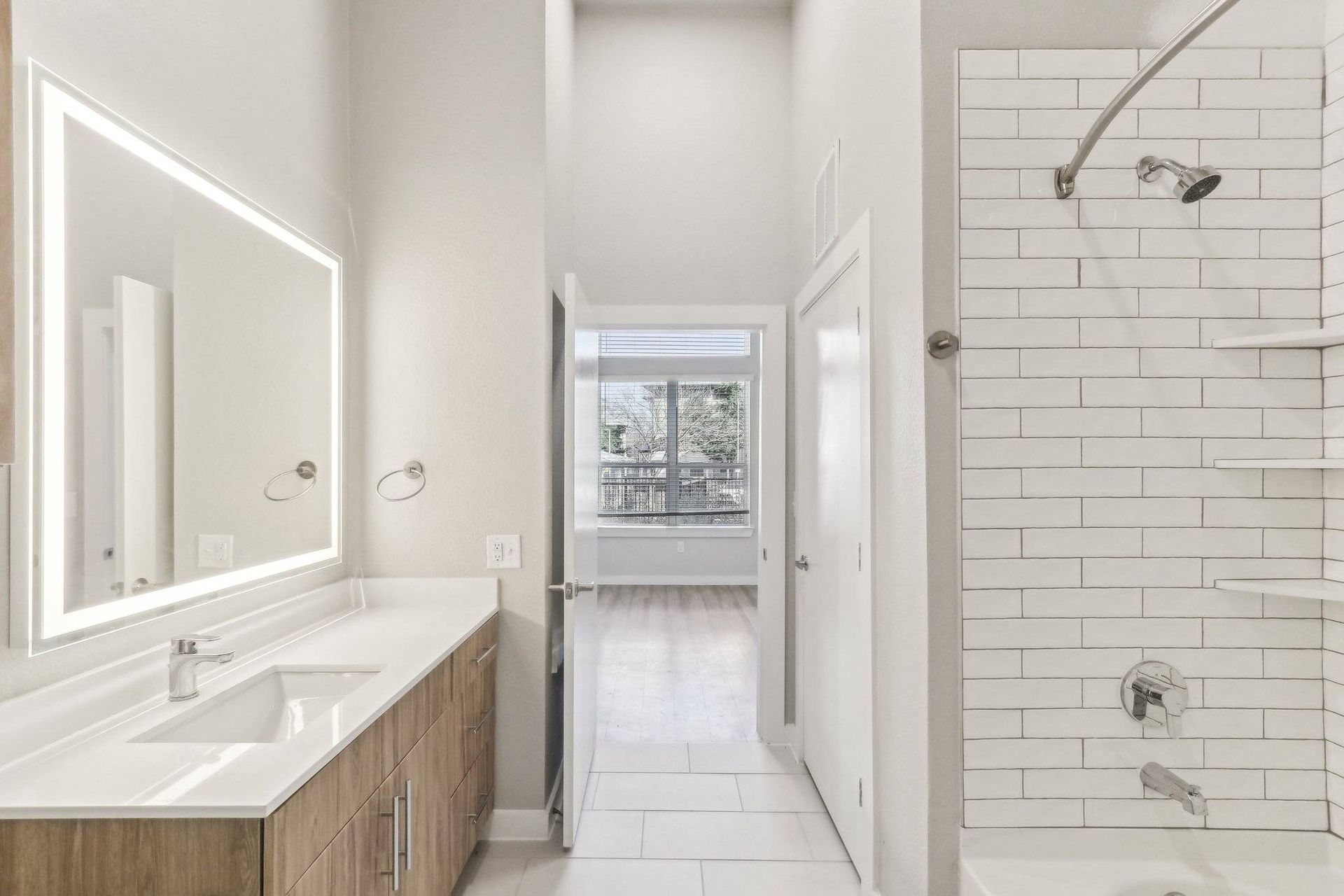 Modern apartment bathroom with a single vanity, backlit mirror, and white subway-tile shower.