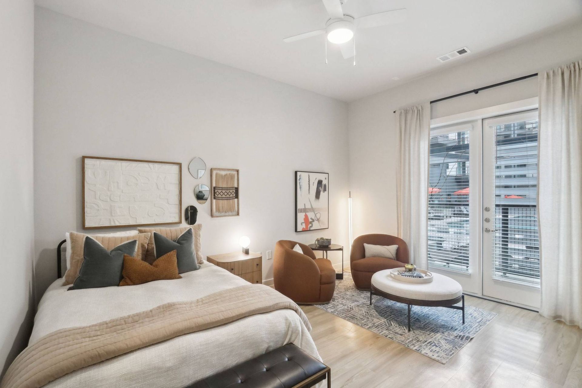 Bedroom and living area with a bed, two orange chairs, ottoman, and a balcony door with white curtains.