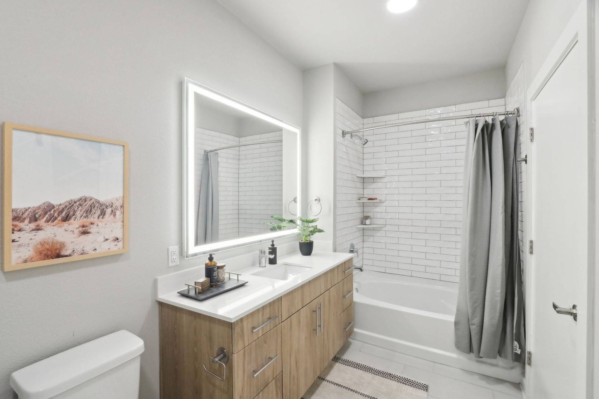 Modern apartment bathroom with a long white vanity, backlit mirror, and subway-tile shower.