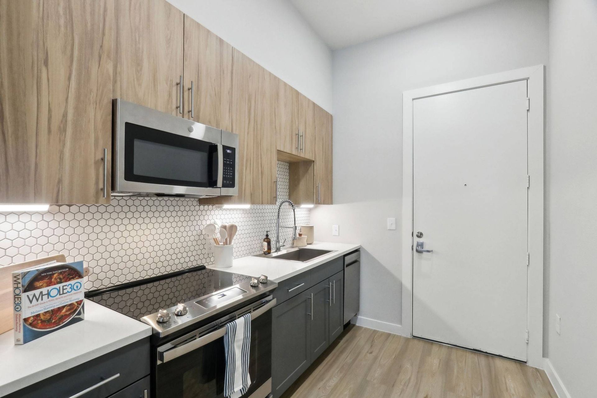Modern apartment kitchen with wood upper cabinets, stainless steel appliances, and a white hex tile backsplash.
