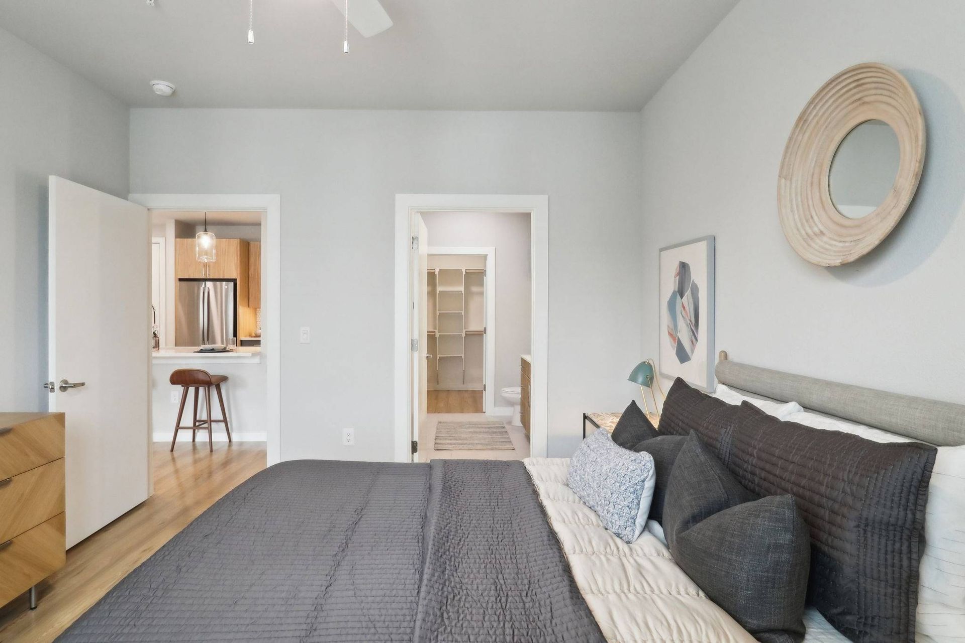 Bedroom with gray bedding, round wall mirror, dresser, and open doorway to a kitchen.