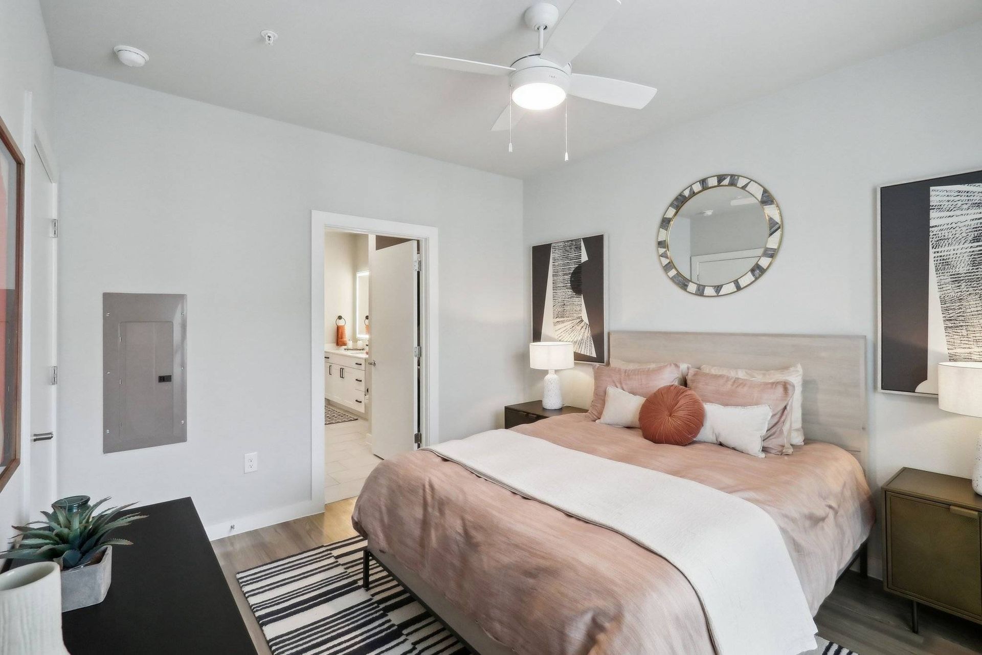 Bedroom with beige bedding, round mirror, two nightstands, and a ceiling fan.