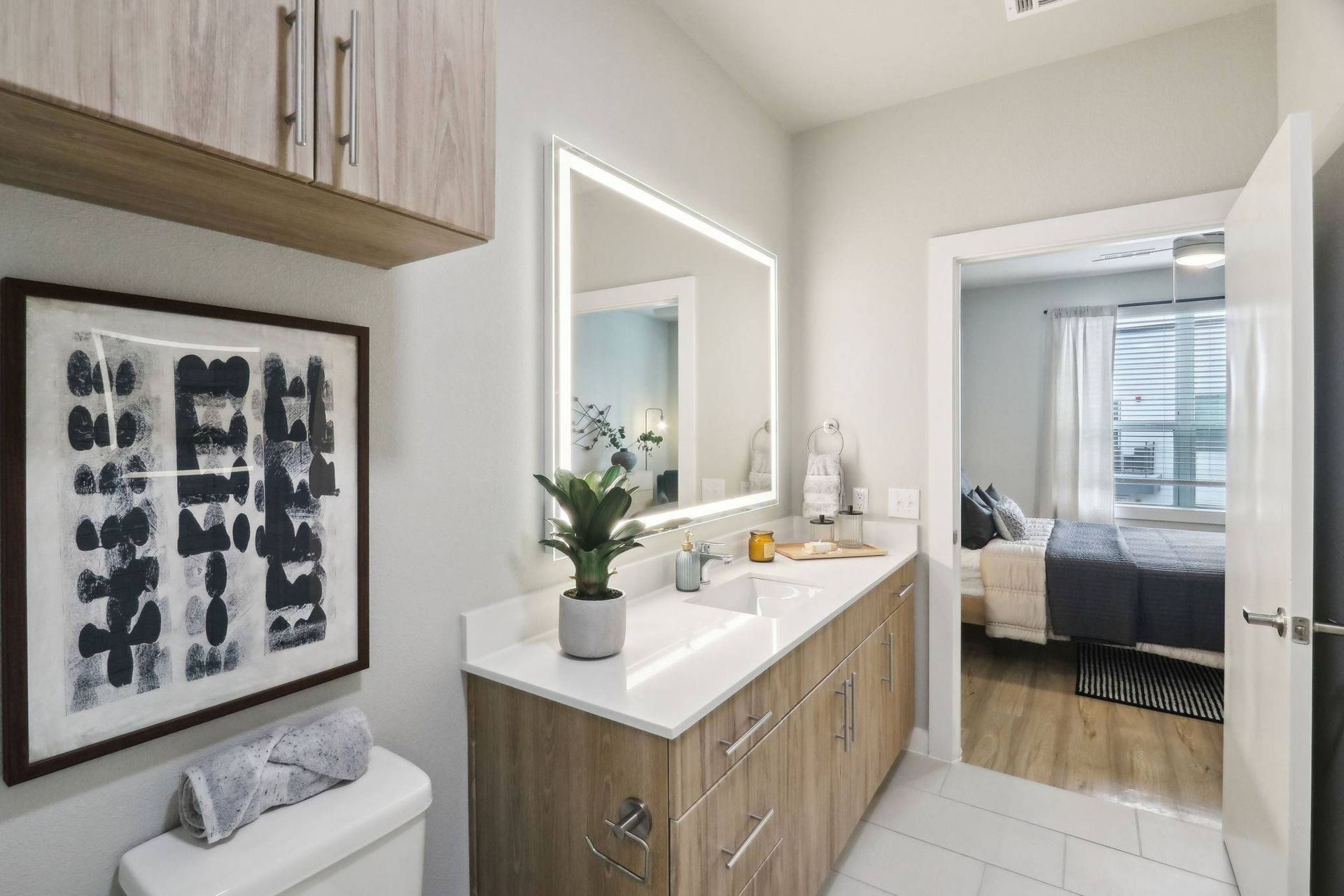 Bathroom vanity with dual sinks, illuminated mirror, and doorway to a bedroom.