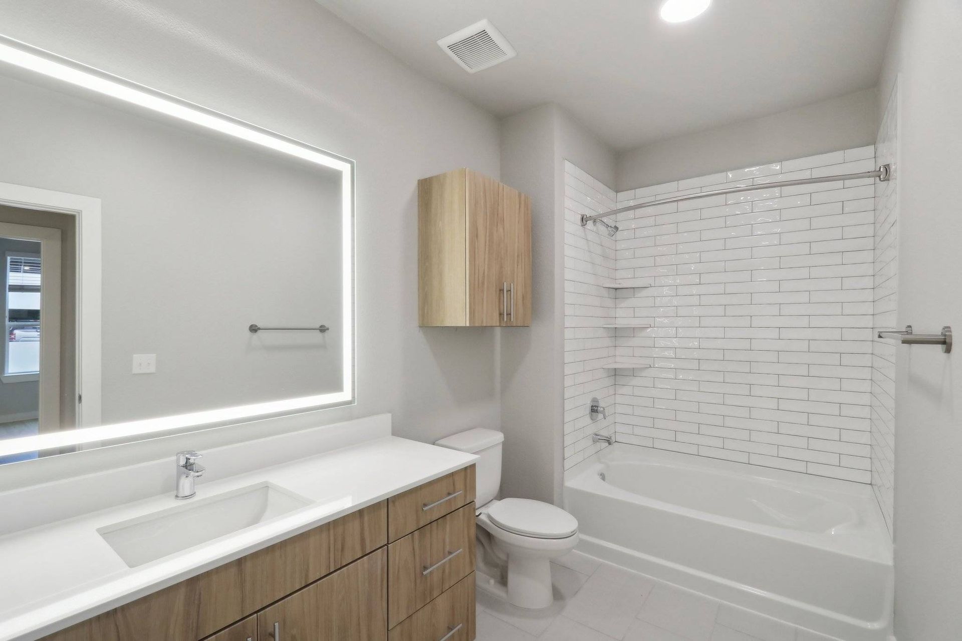 Modern bathroom with white vanity, backlit mirror, toilet, and tub/shower with subway tile.