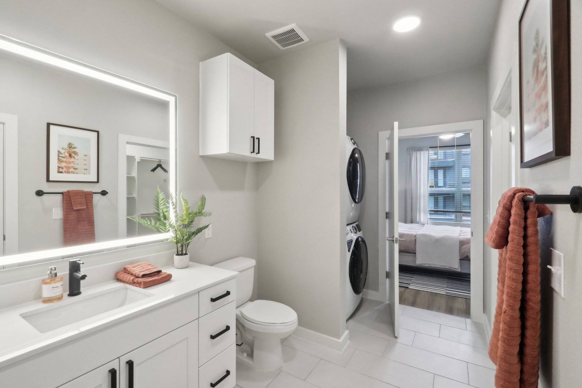 Modern white bathroom with backlit mirror, vanity, and stacked washer/dryer visible through doorway.