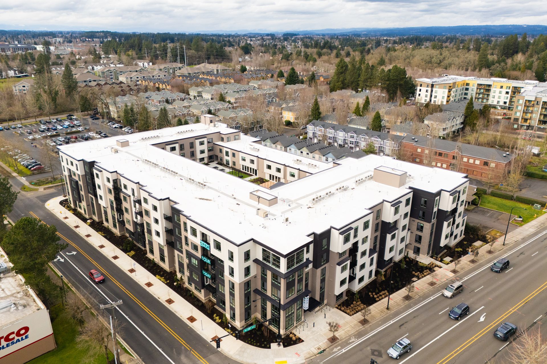 Aerial view of a U-shaped apartment building at a road intersection near a parking lot with trees and suburban housing.