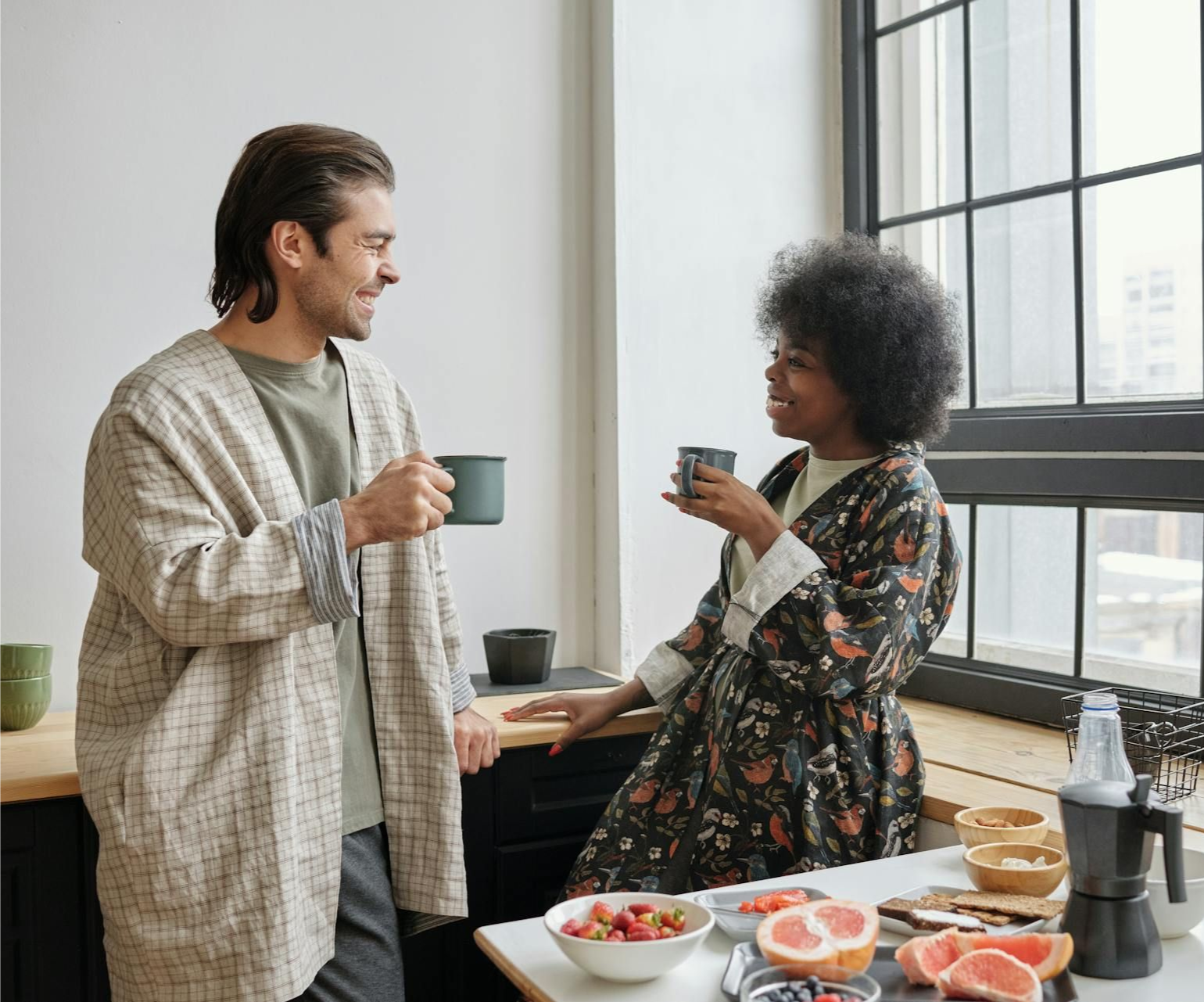 Couple in bathrobes, drinking coffee, chatting near a window, with fruit on counter.