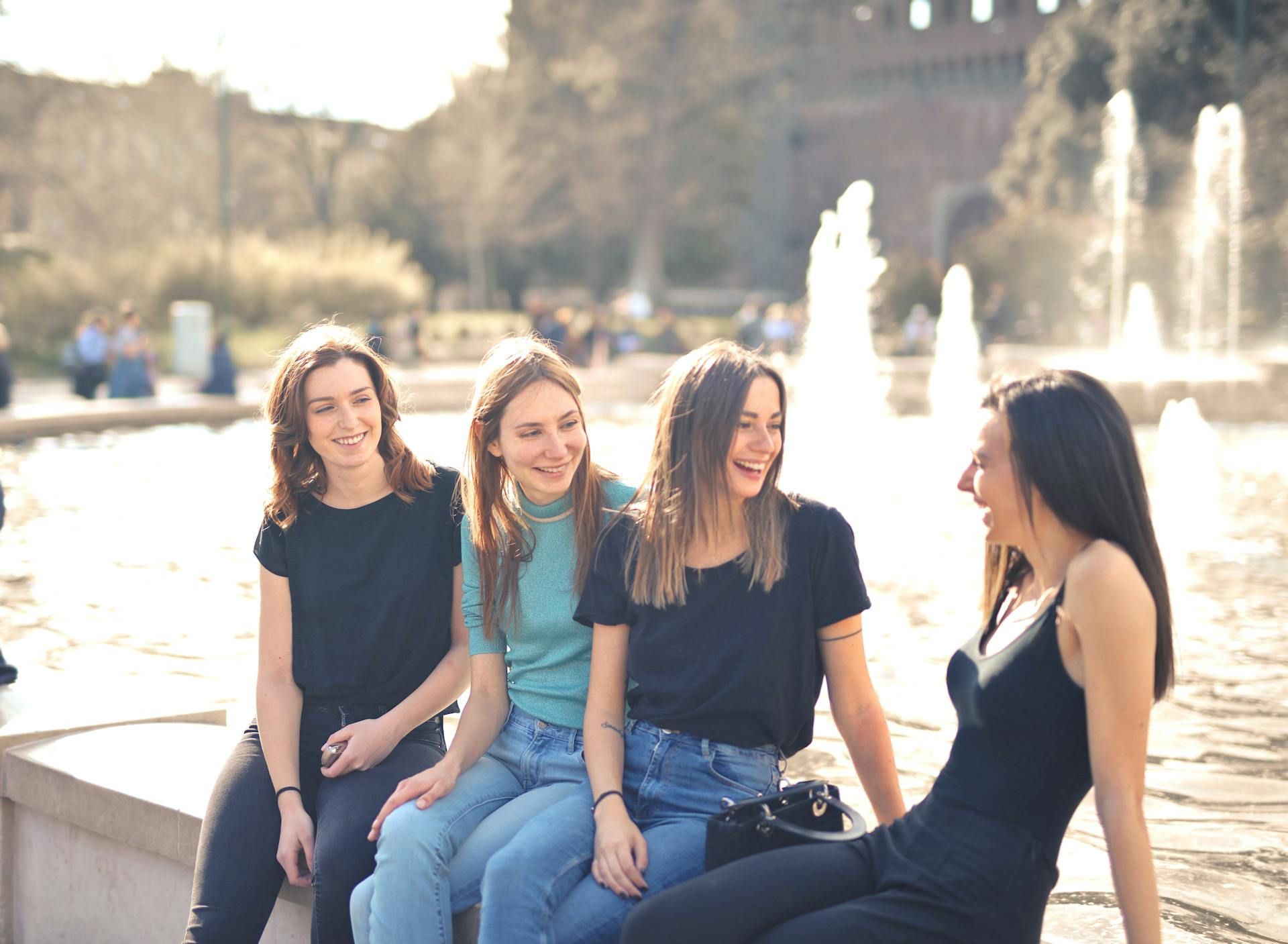 Four women laughing, sitting on a wall in front of a fountain. Sunny outdoor scene.