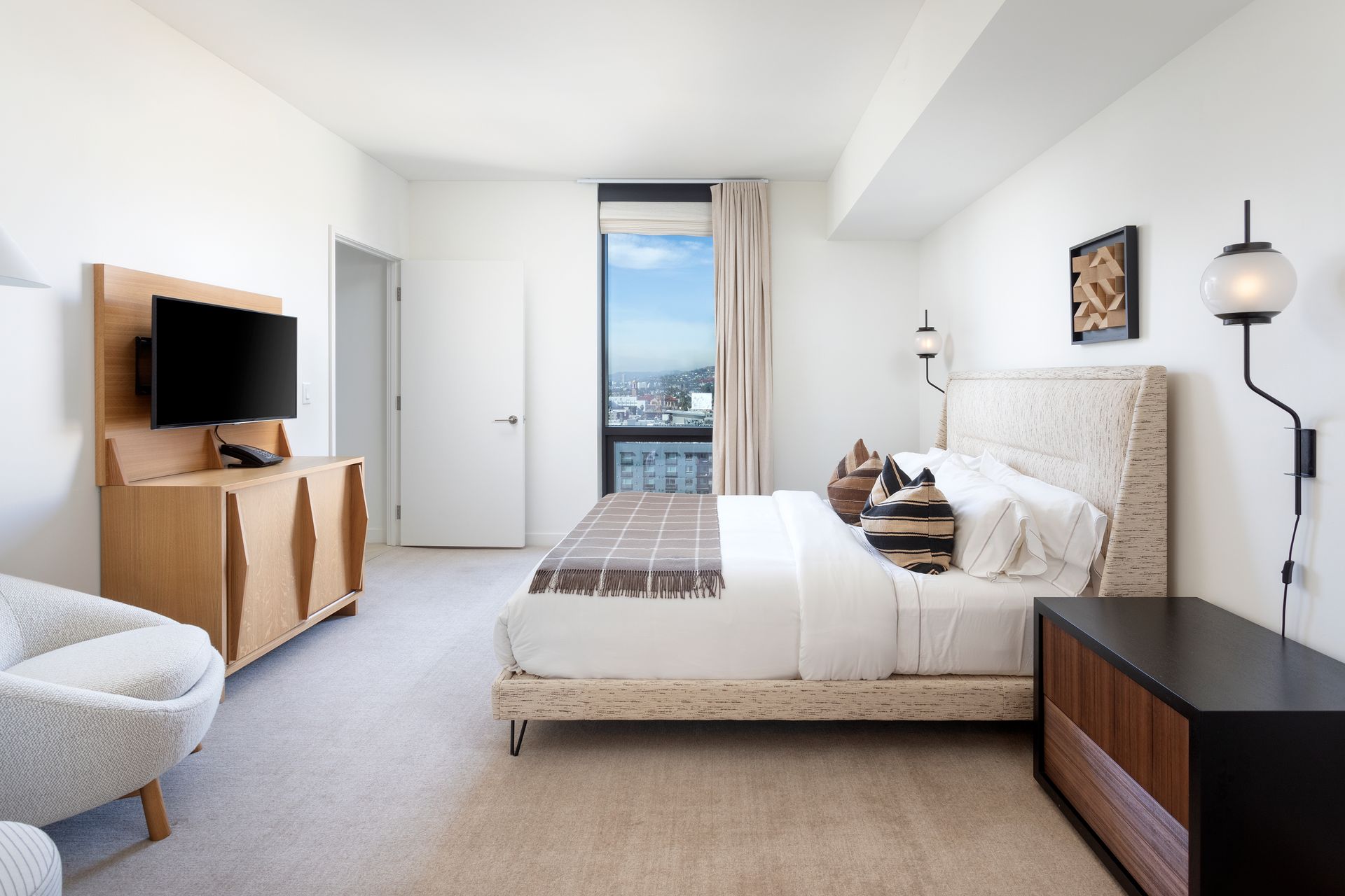 Bright modern bedroom with a bed, TV, dresser, and a window view of the city.
