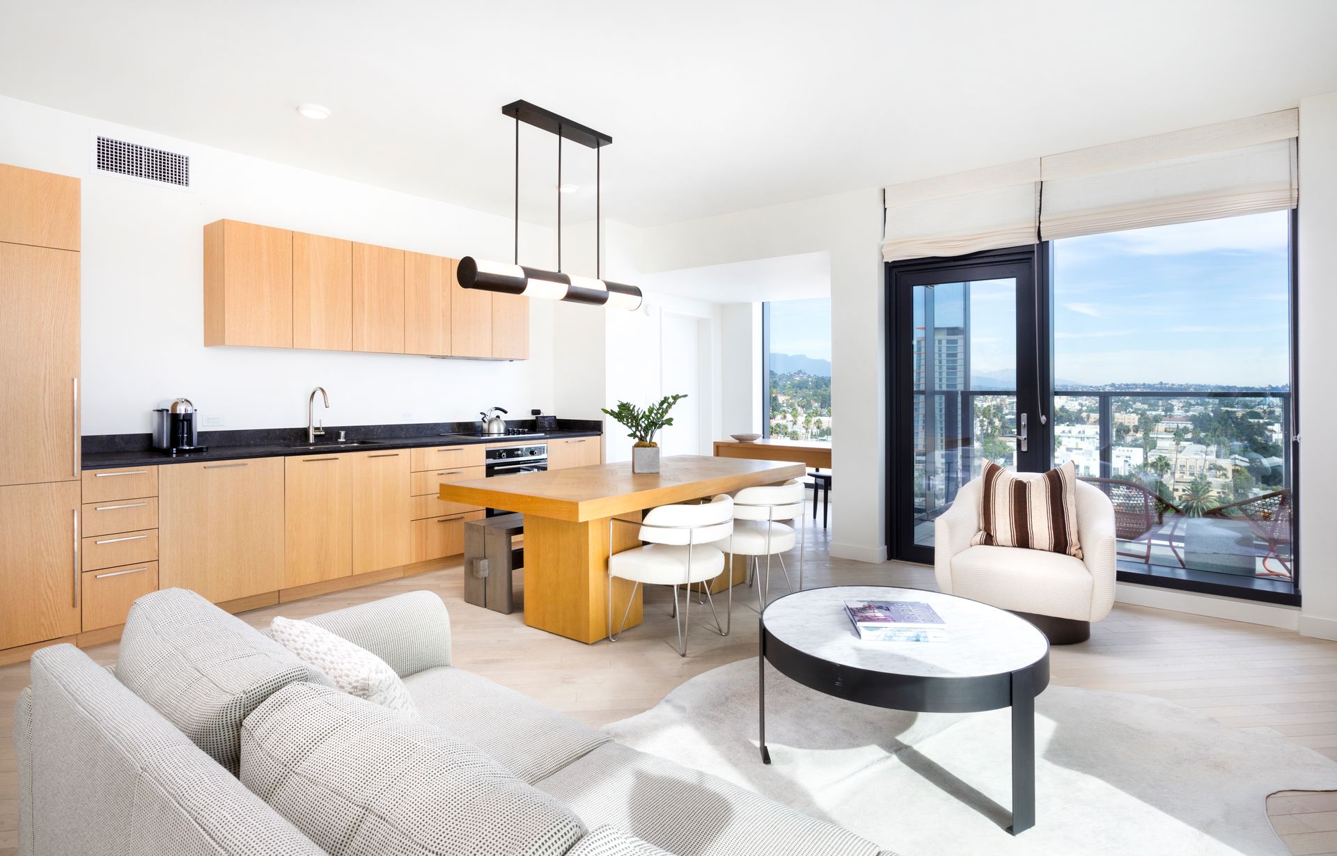 Bright modern living-dining room with wood cabinets, white sofa, and floor-to-ceiling windows overlooking a city view