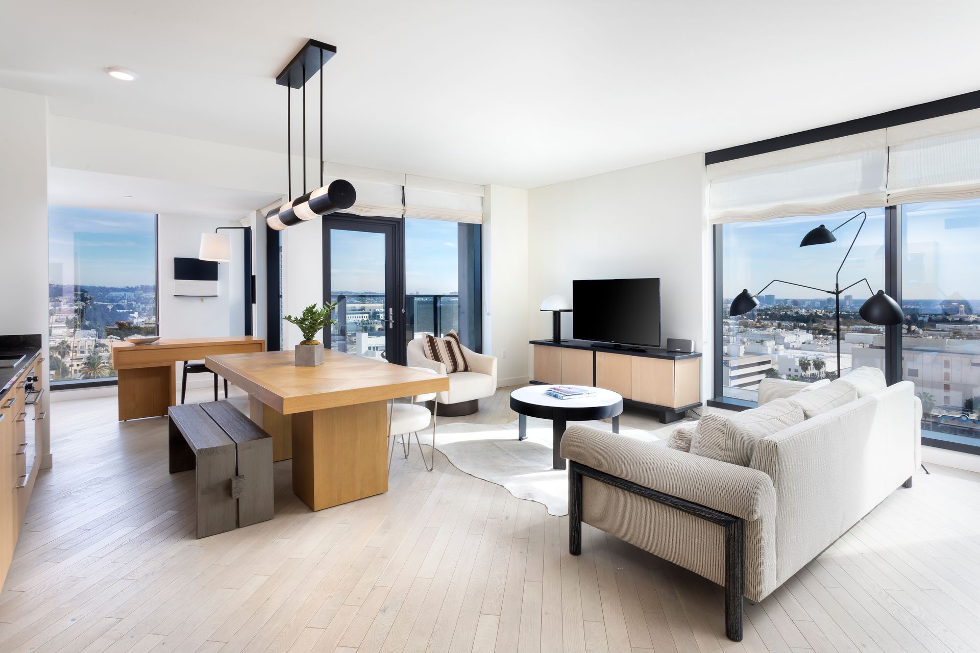 Bright modern open-plan living room with sofa, dining table, kitchen island, TV, and city-view windows