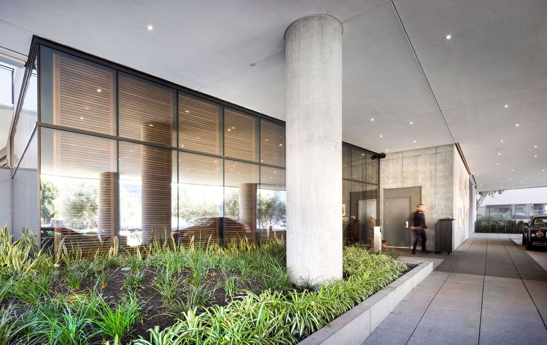 Modern building entrance with glass walls, concrete columns, landscaped plants, and a person near the doorway