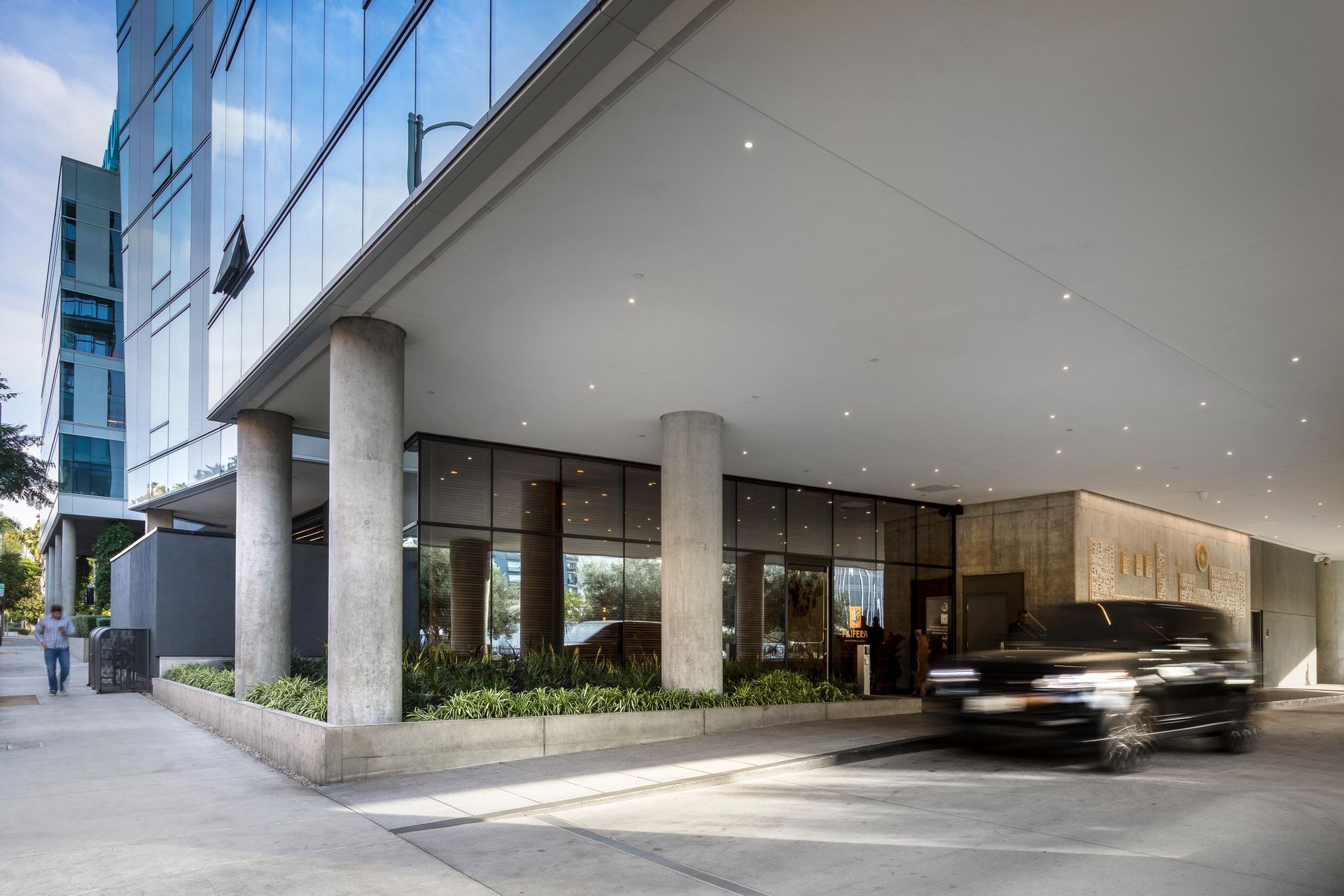 Modern building entrance with glass windows, concrete columns, and a car driving by under an overhang