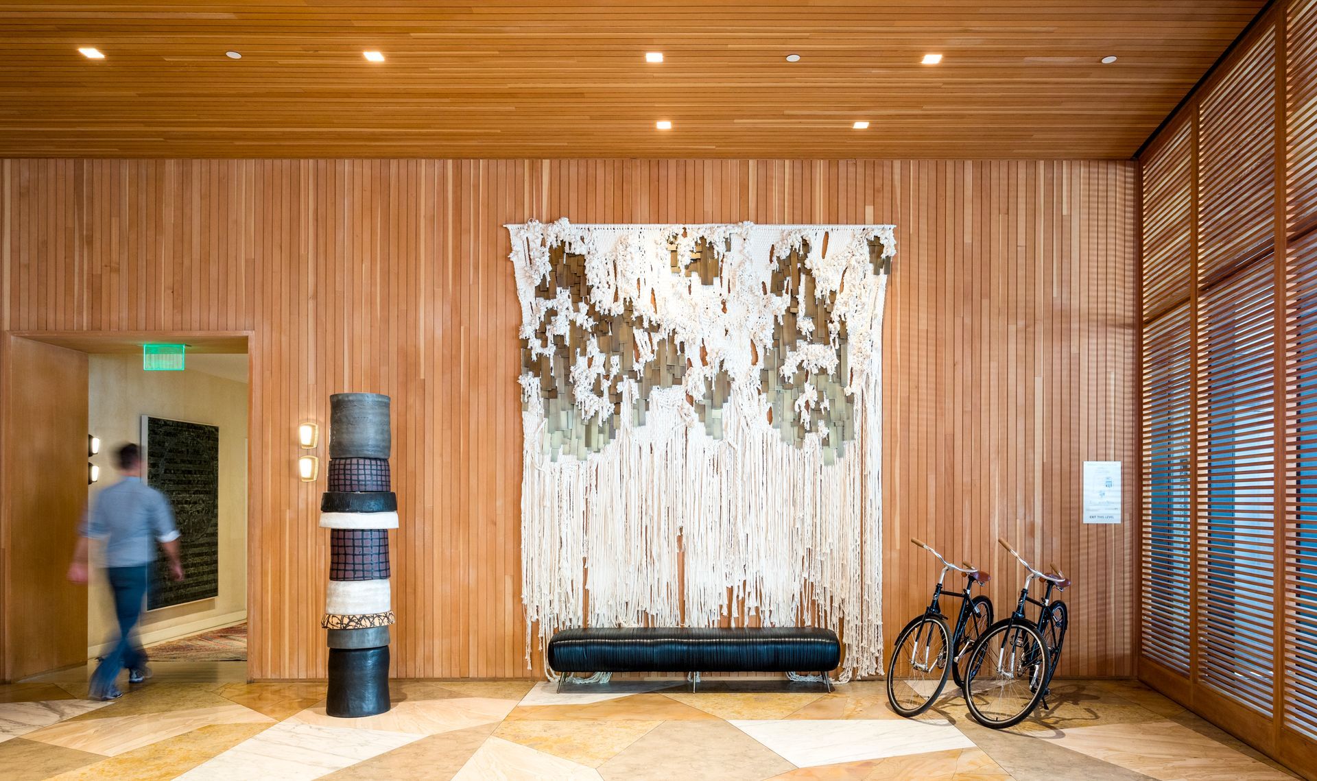 Wood-paneled lobby with abstract wall art, bench, bicycles, and a blurred person walking by the entrance
