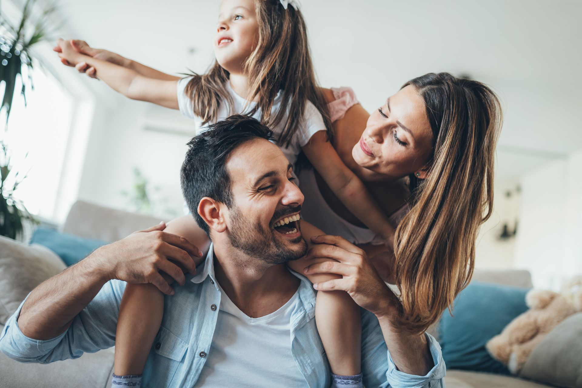 Three people laughing together on a couch in a bright living room, with one child on an adult’s shoulders.