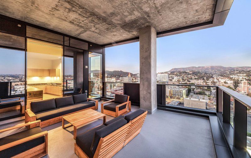 Modern apartment balcony with wooden lounge chairs, gray seating, and city views at sunset