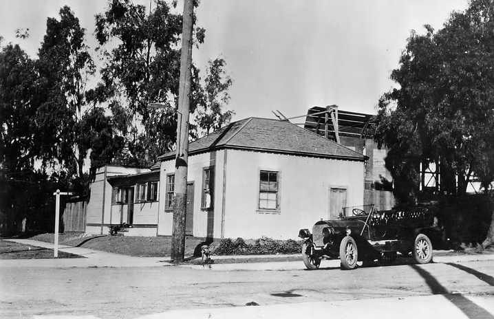 Black-and-white photo of a small house with trees and a vintage car parked at the curb.