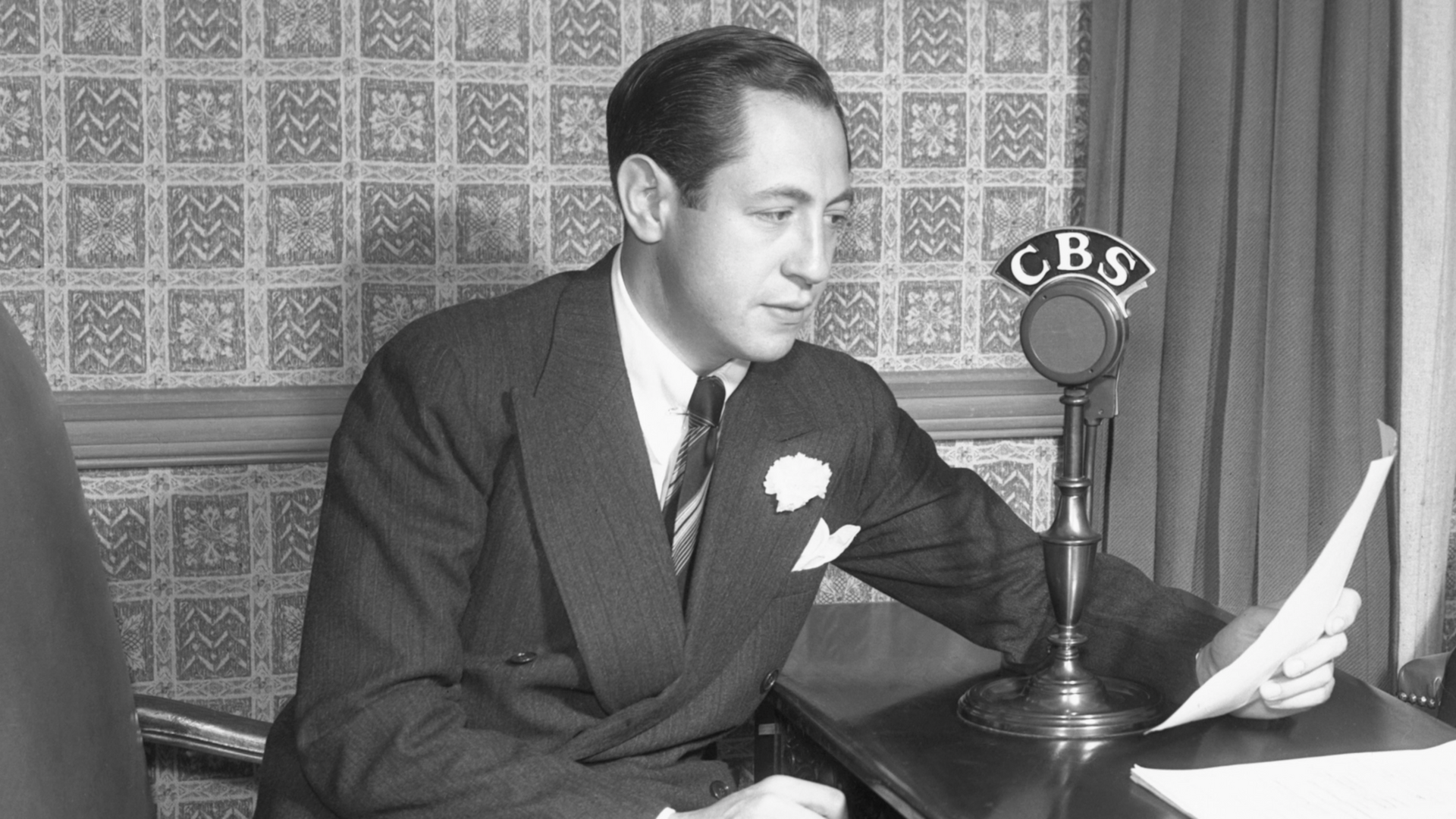 Man seated at a desk speaking into a CBS radio microphone while reading papers