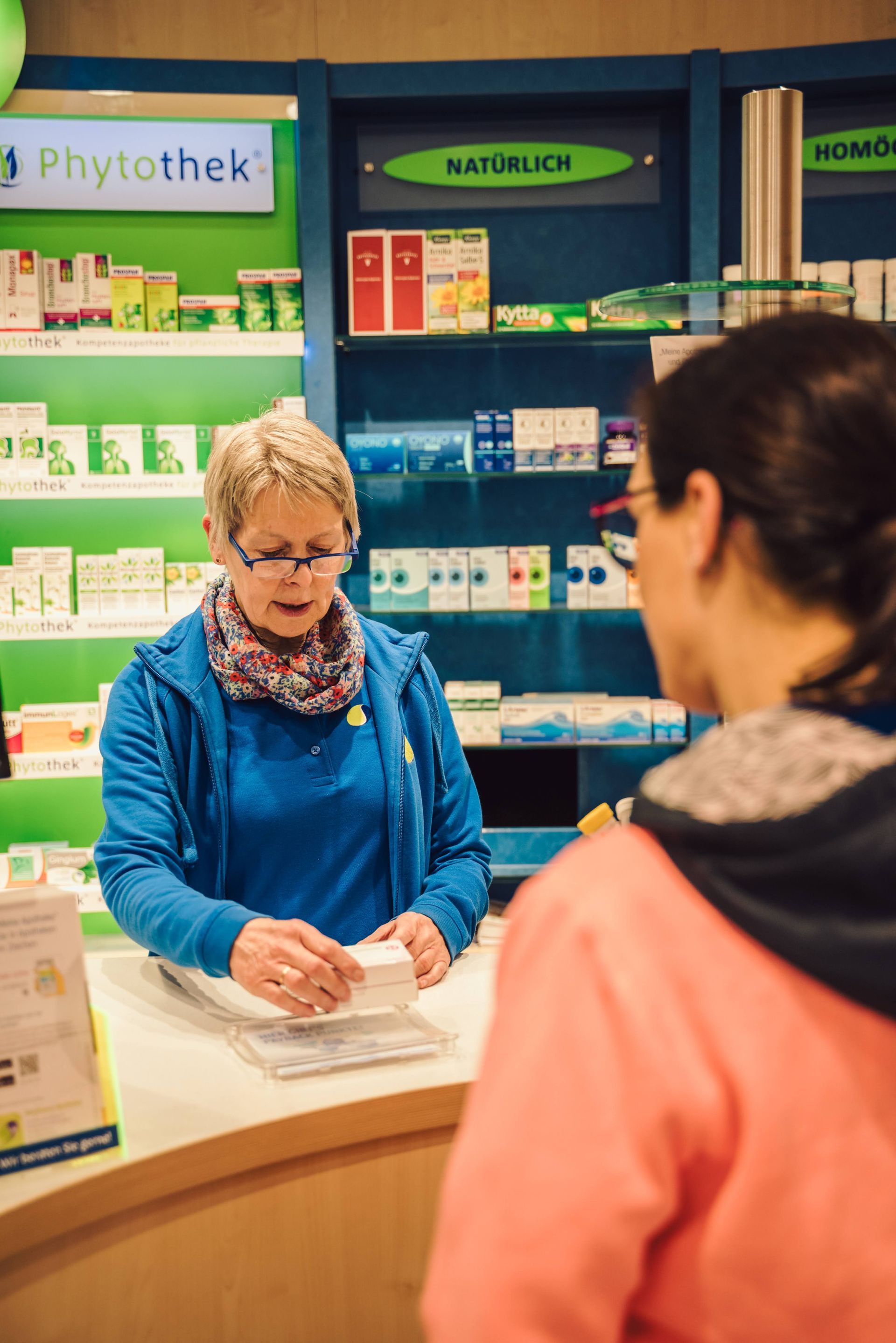 Eine Frau sitzt an einem Tresen in einer Apotheke und unterhält sich mit einem Kunden.