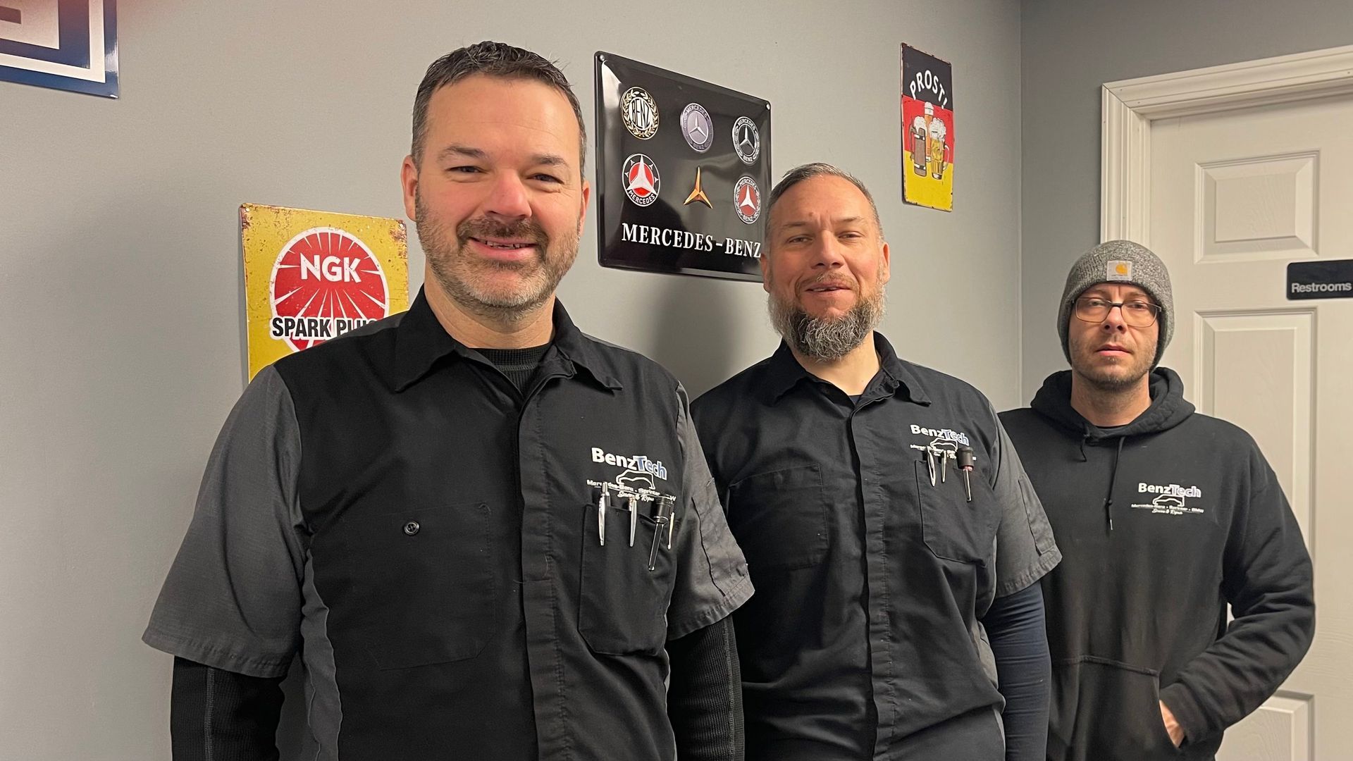 Three auto shop workers in black shirts, smiling in a garage setting, with tools in shirt pockets. | Benz Tech
