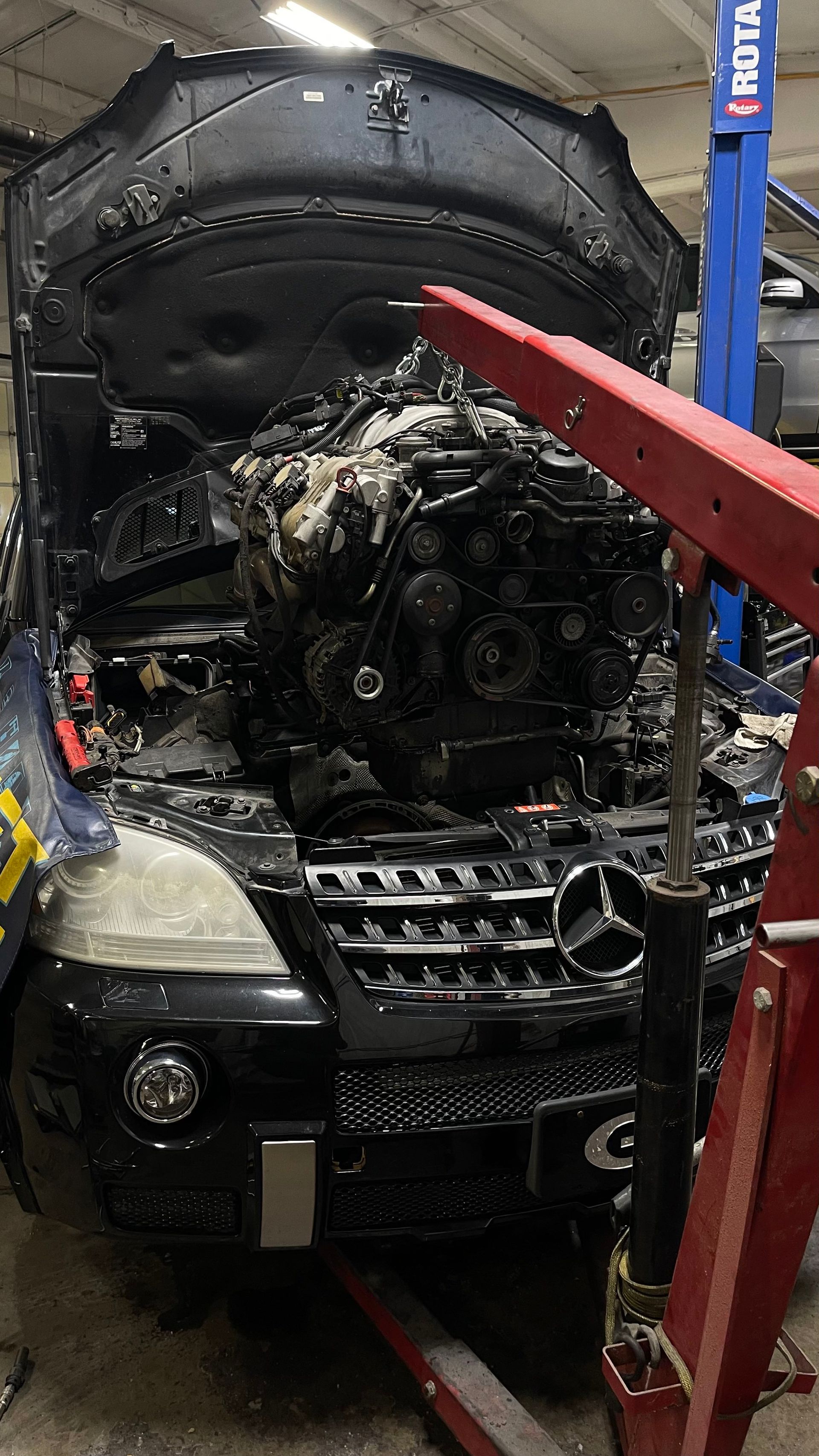 A black Mercedes with its hood open, engine being removed by a red hoist in a repair shop. | Benz Tech