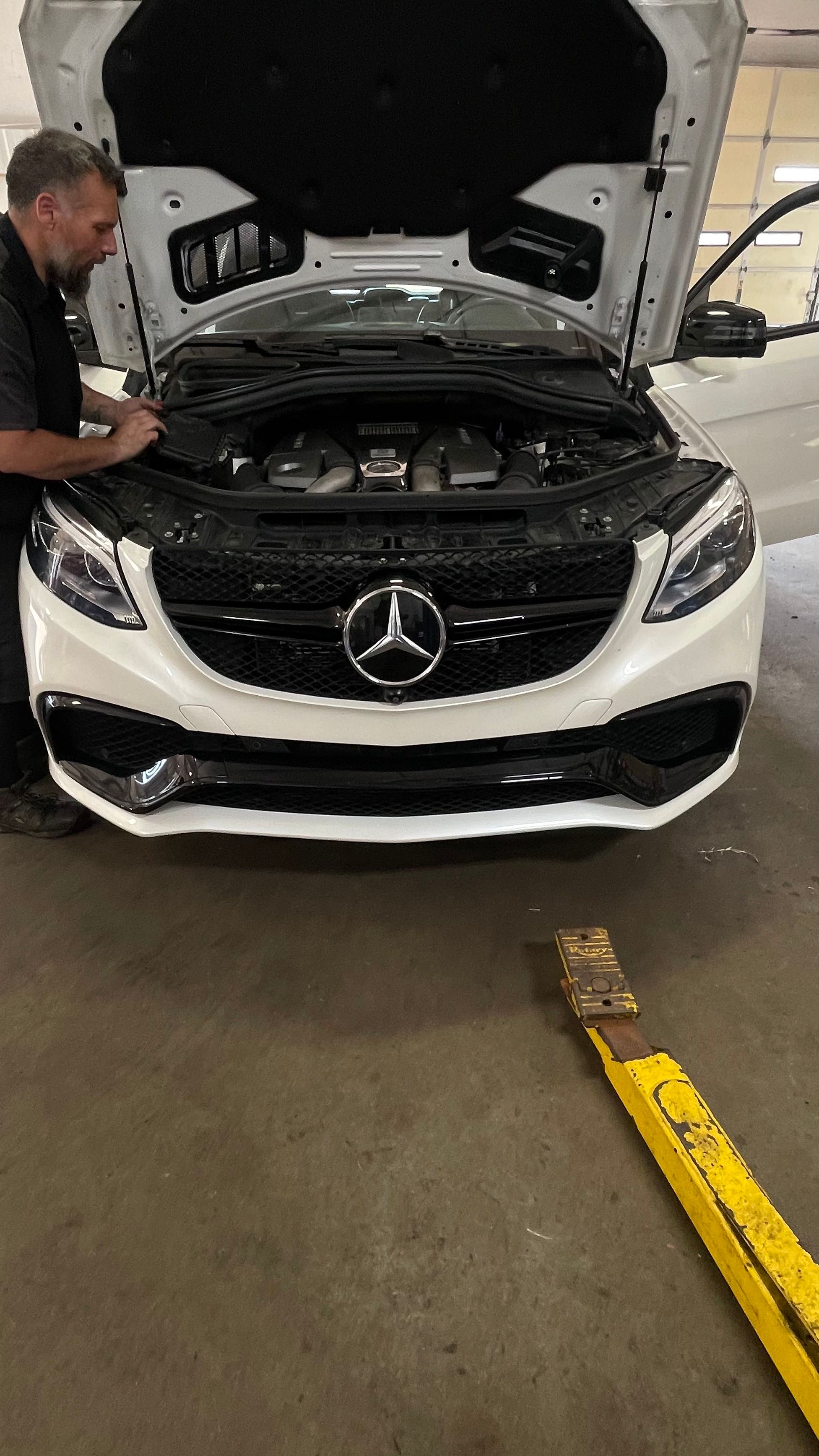 Man inspecting a white Mercedes with open hood in a garage; yellow jack in foreground. | Benz Tech