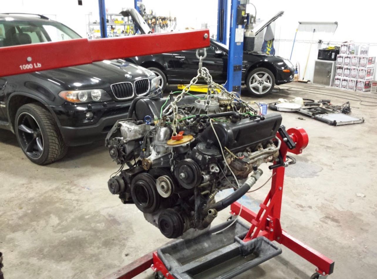 Engine being lifted by a crane in a garage, with a BMW SUV and sedan in the background. | Benz Tech