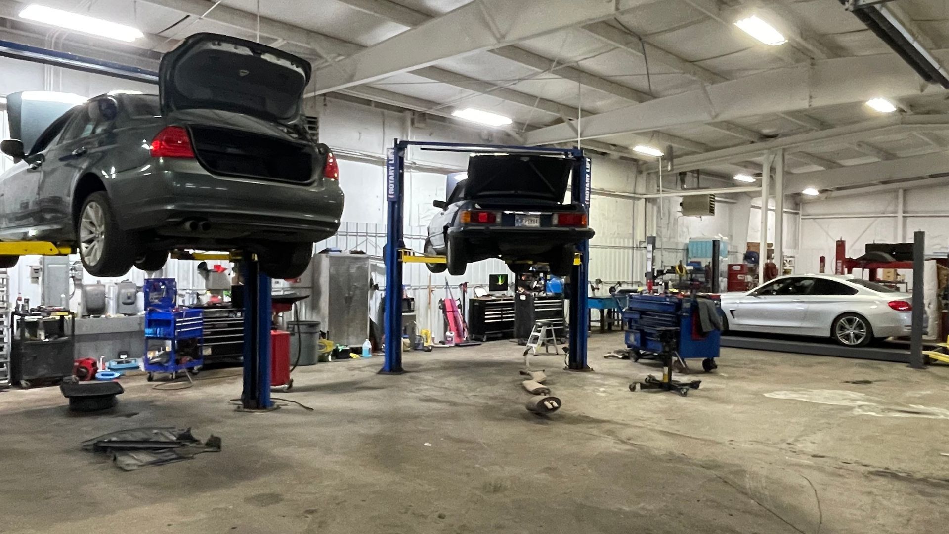 A car repair shop with two cars raised on lifts. One is gray, the other is black. Various tools are scattered about. | Benz Tech