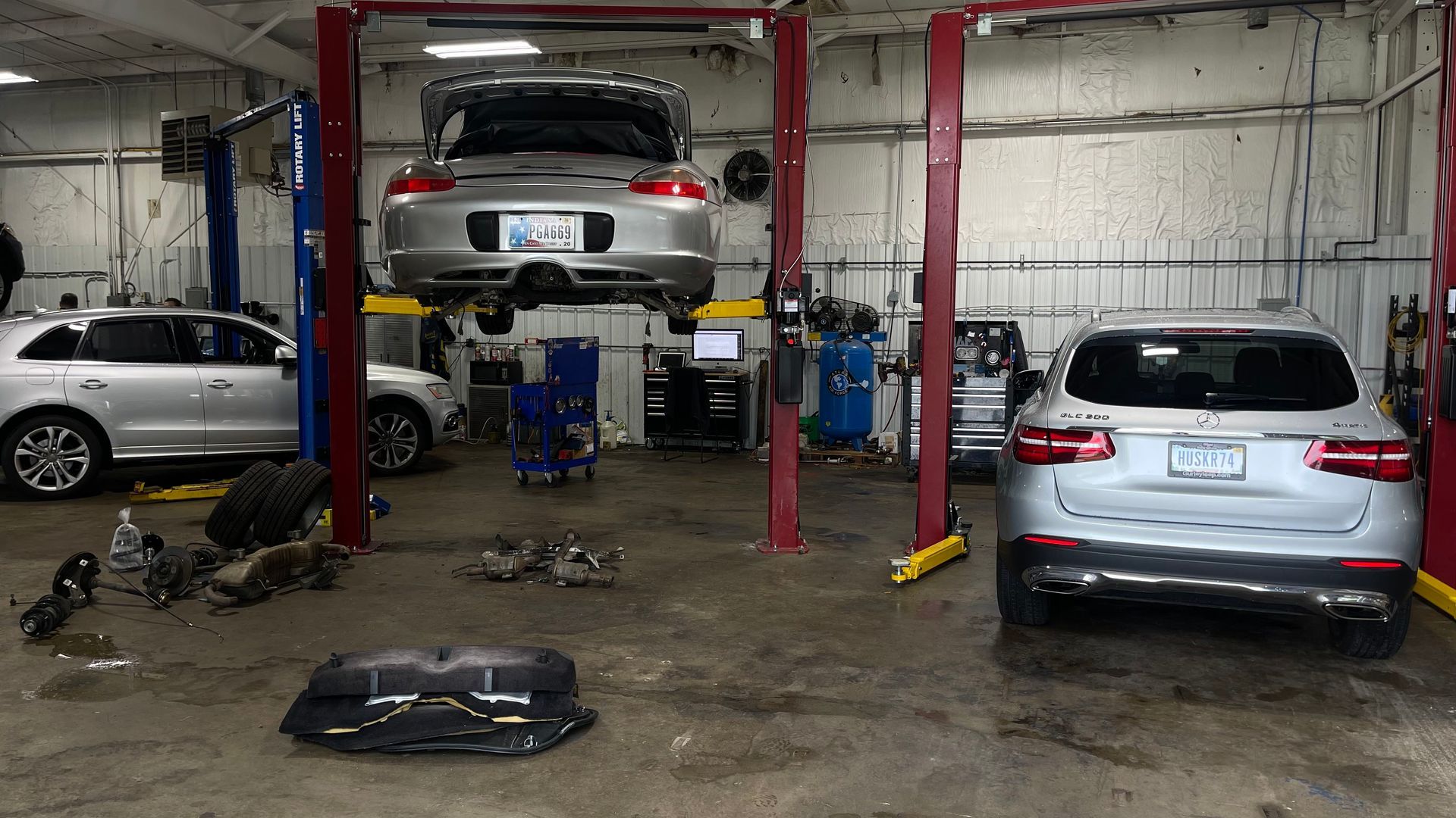 A silver Porsche on a car lift in a garage, with other cars, tools, and parts scattered around. | Benz Tech