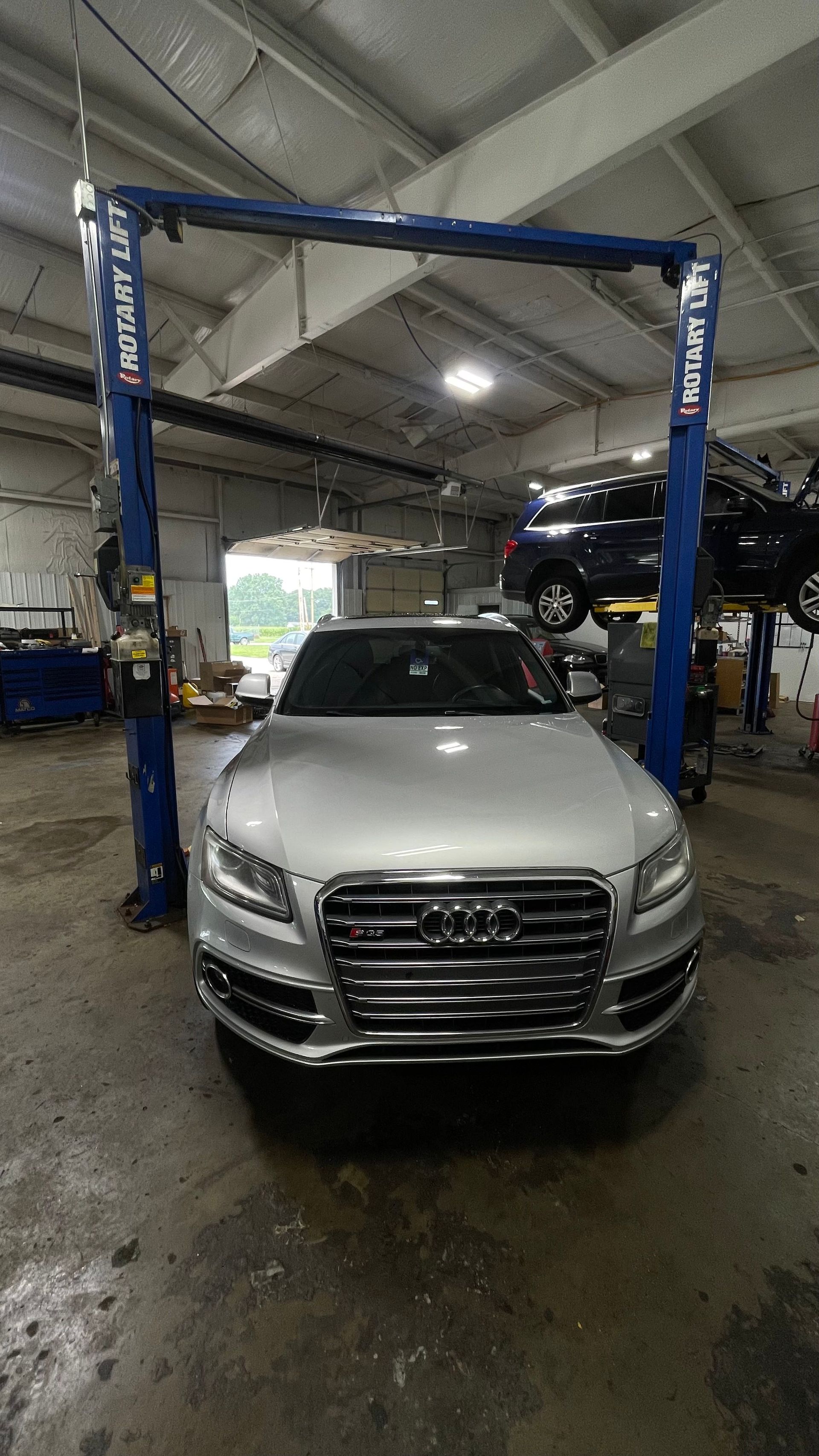 Silver Audi SUV in a repair shop, facing forward, with another car lifted above. | Benz Tech