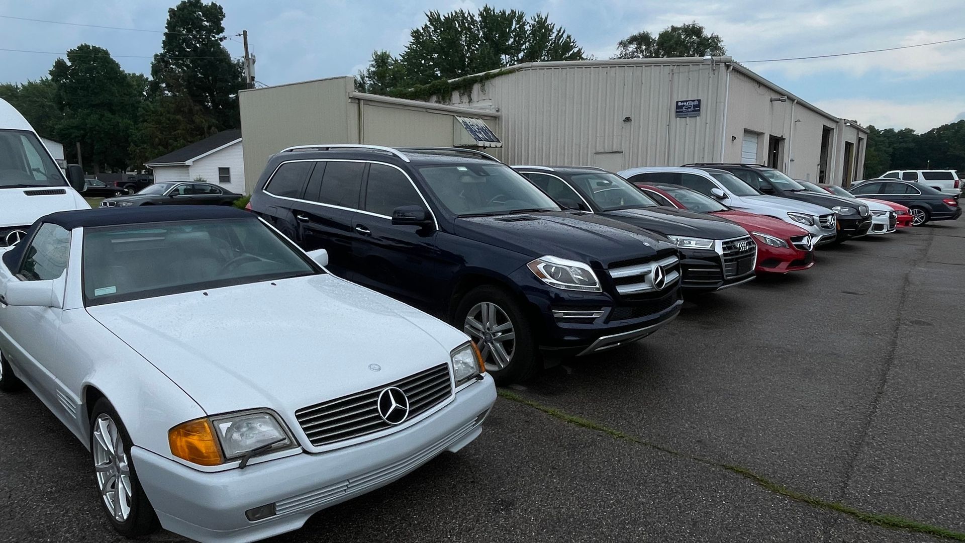 A row of cars, mostly Mercedes-Benz, parked in front of a building, under a cloudy sky. | Benz Tech