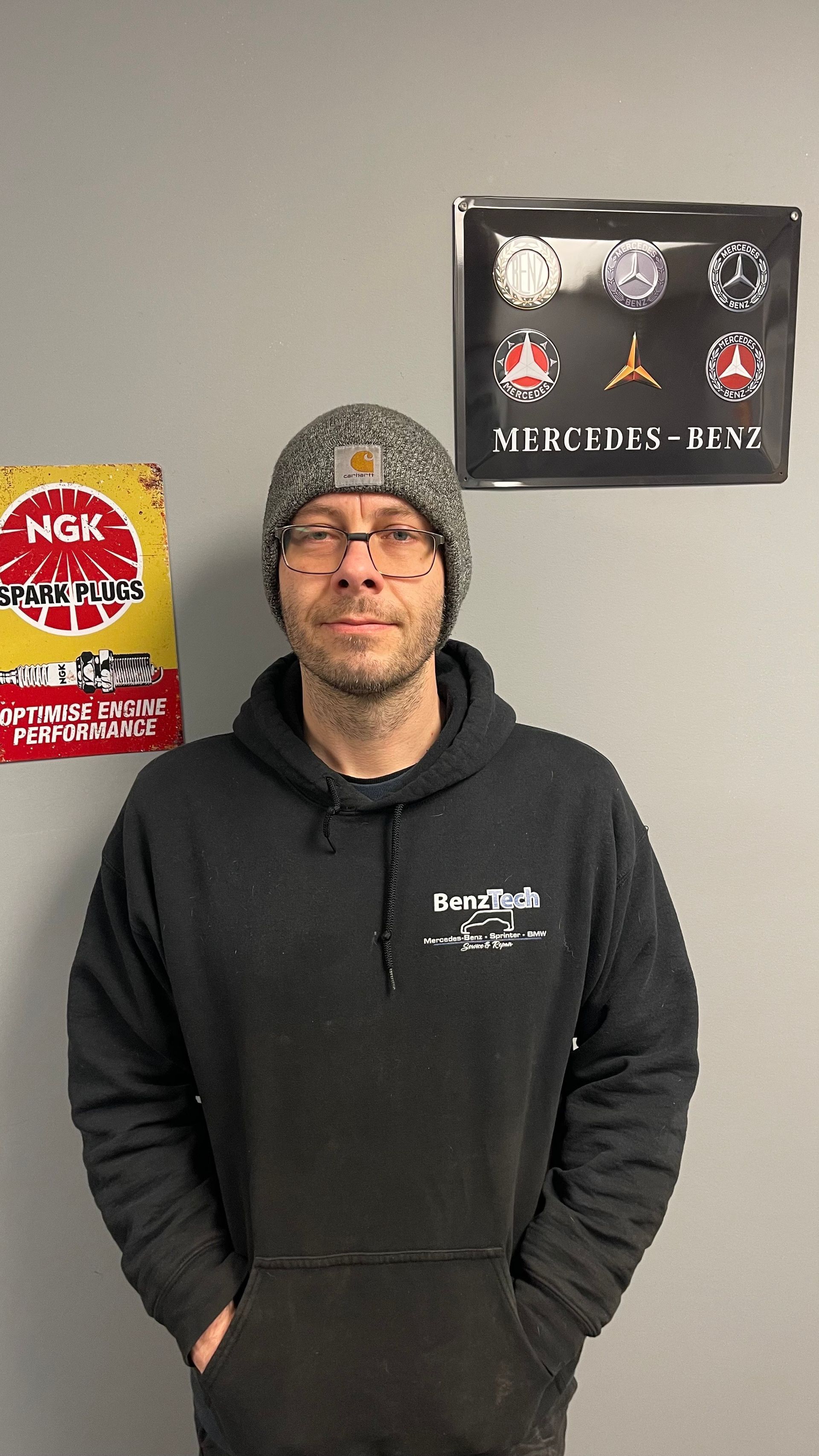 Man in black hoodie and beanie, hands in pockets, in front of garage signs. | Benz Tech