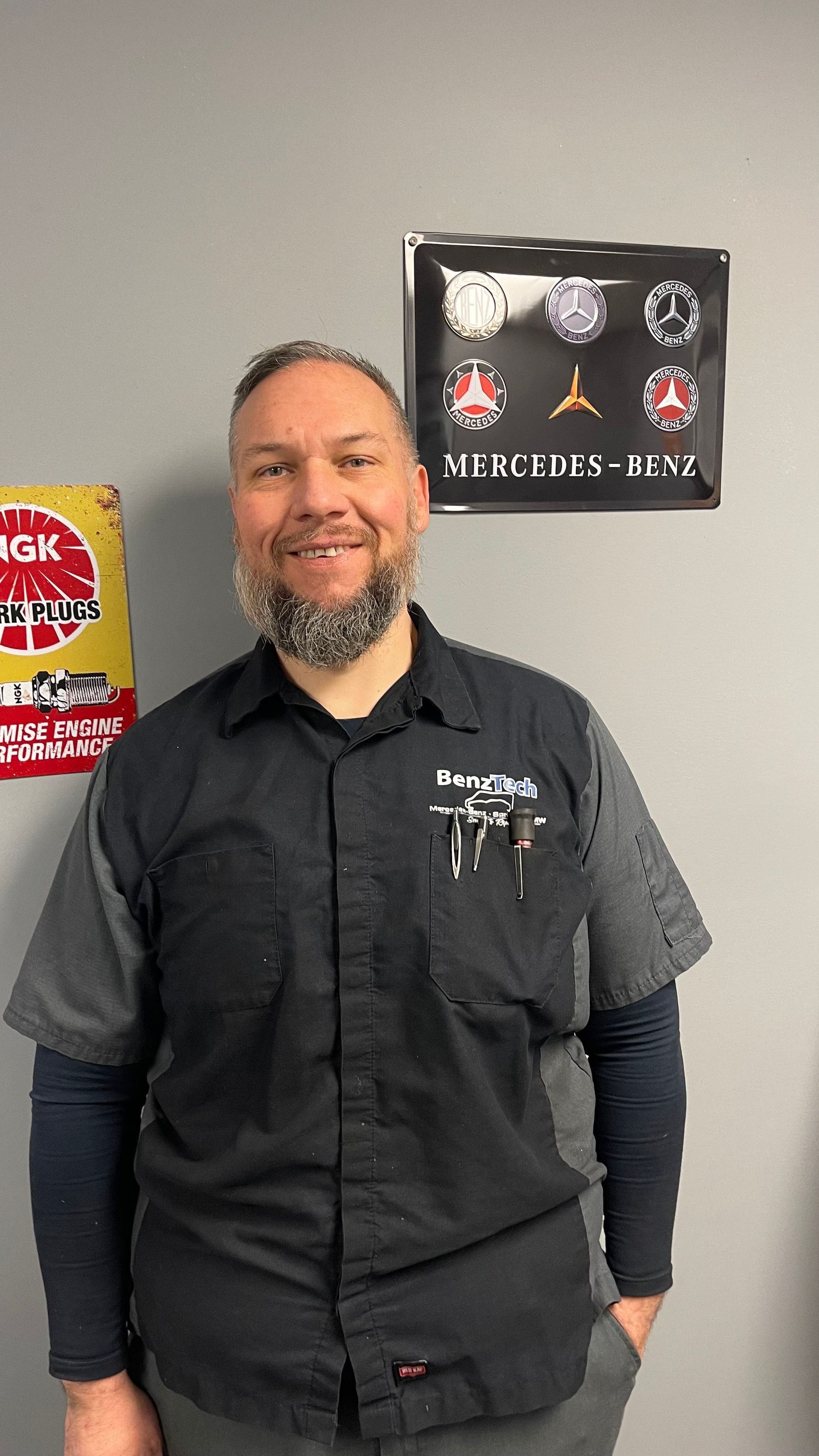 Mechanic in gray and black uniform smiles, standing next to a Mercedes-Benz sign and spark plug poster. | Benz Tech