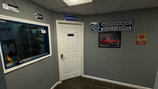 Inside a Mercedes-Benz service shop. A door and window are on a gray wall, with Mercedes-Benz signs and a classic car photo. | Benz Tech