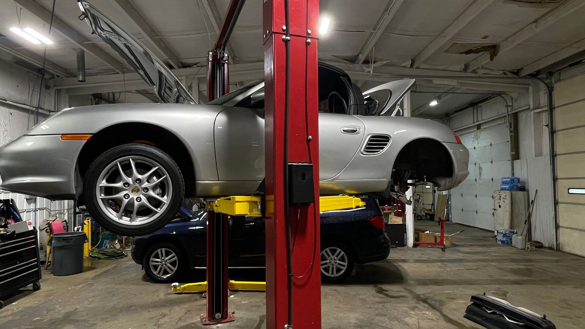 Silver Porsche Boxster on a lift in a garage with a blue car beneath it. | Benz Tech