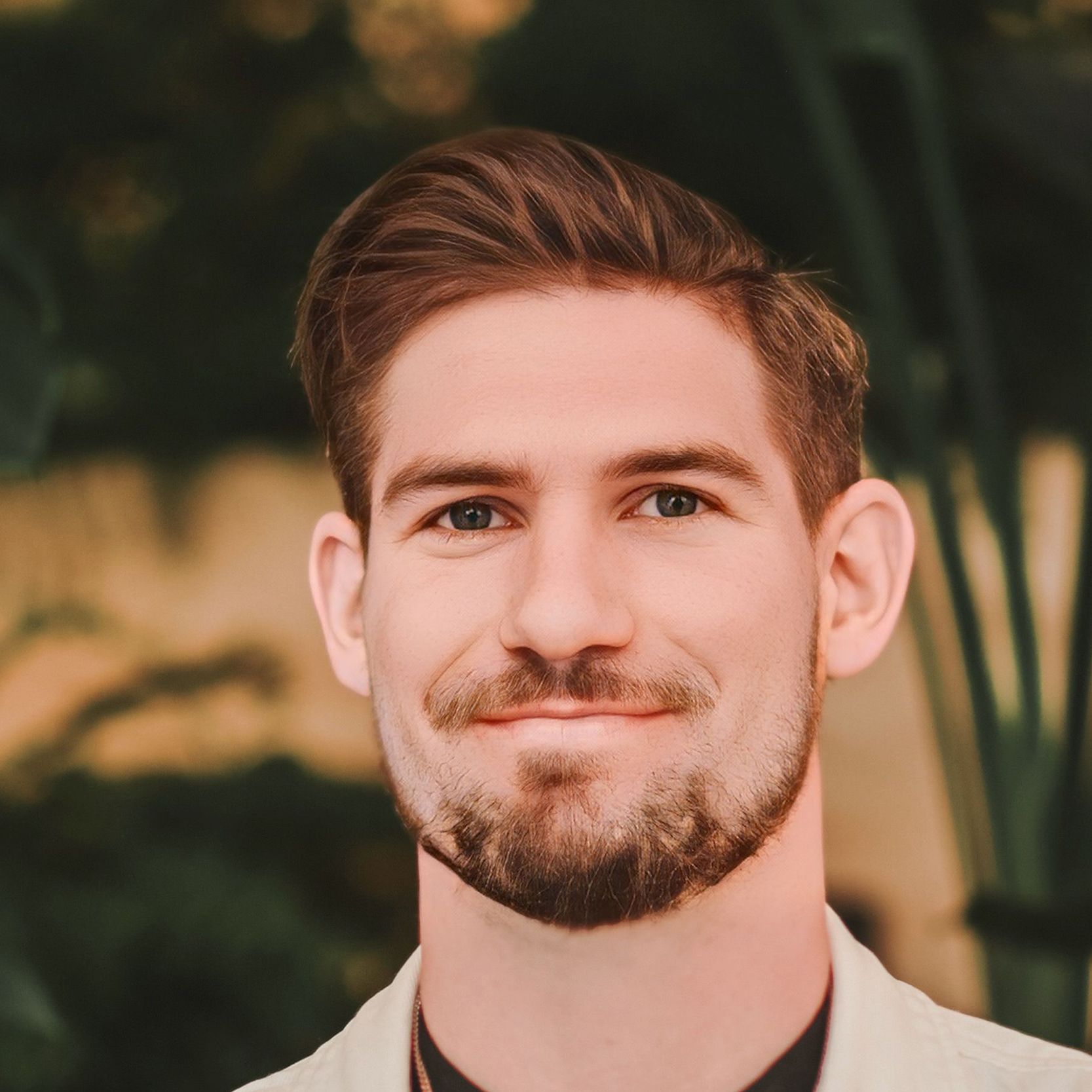 Woman with short blonde hair smiles, wearing blue top and turquoise necklace against a blurred outdoor backdrop.