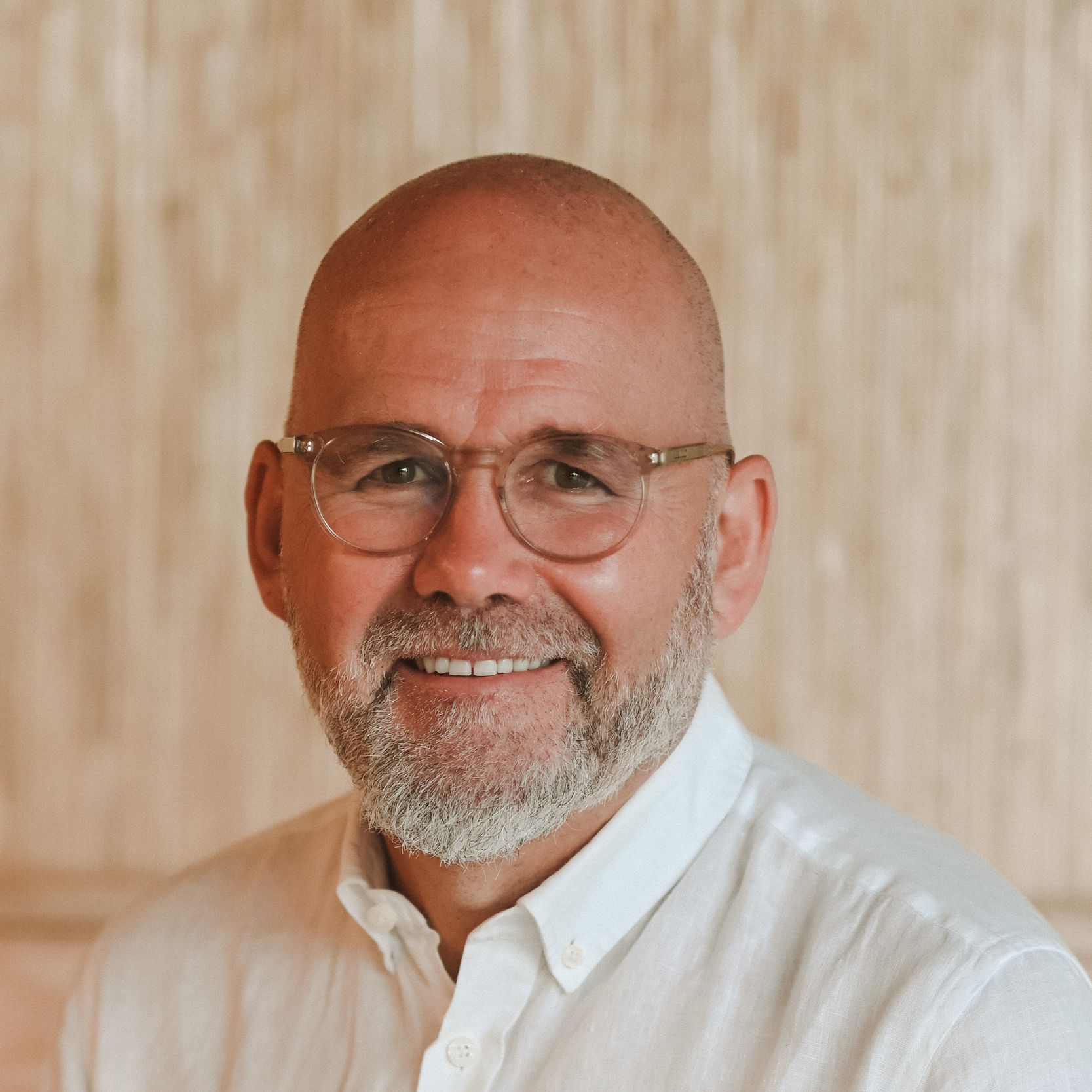 Bald man with glasses smiles, wearing a white shirt; light-colored wall background.